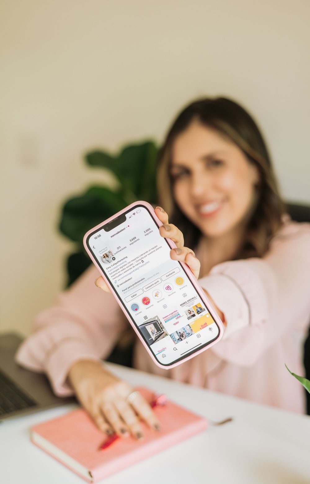Someone holding up a smartphone displaying a social media app while sitting at desk with pink notebook and houseplant.