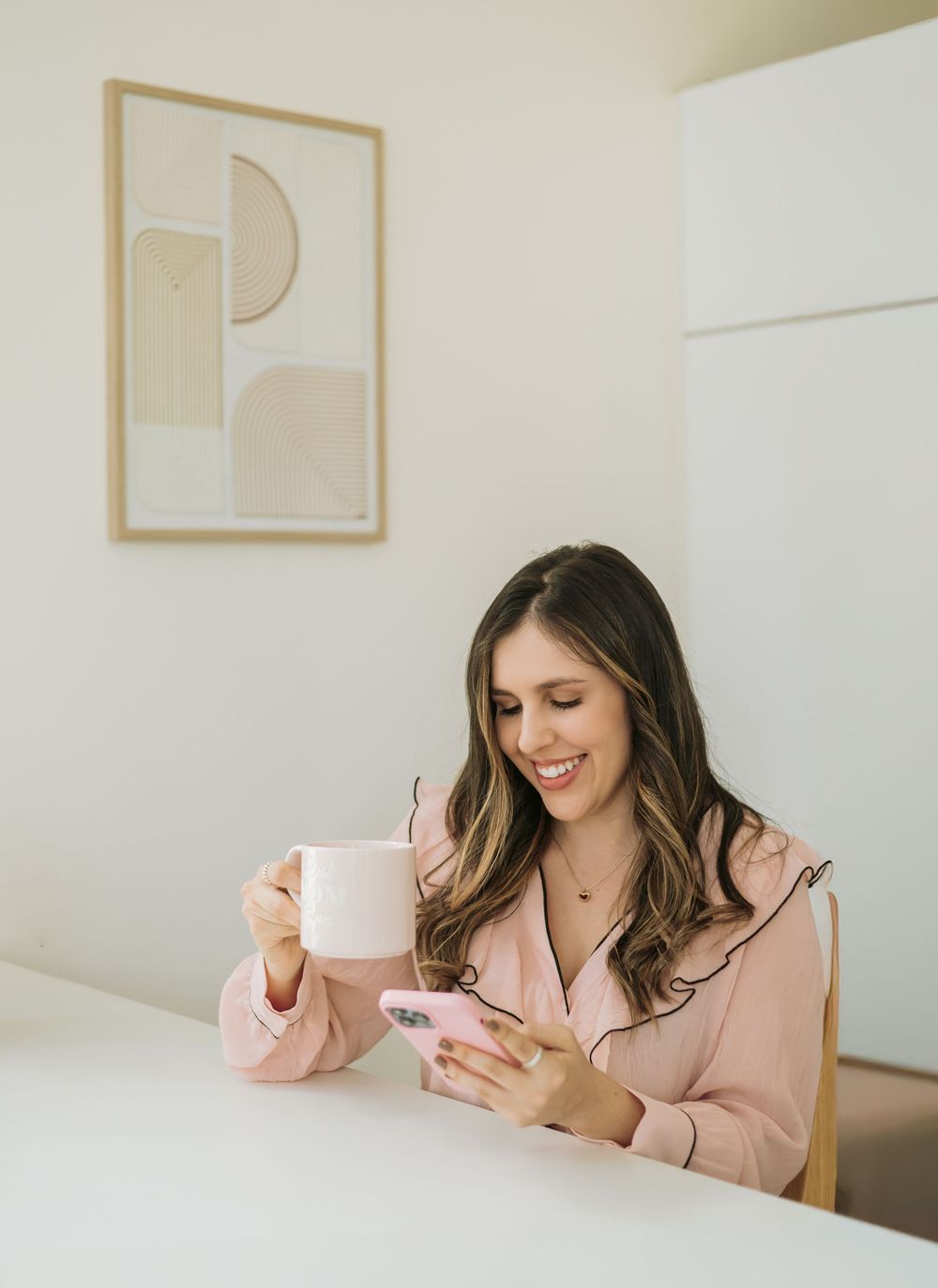 A person in a pink blazer smiles while holding a coffee cup and checking their phone at a white desk.