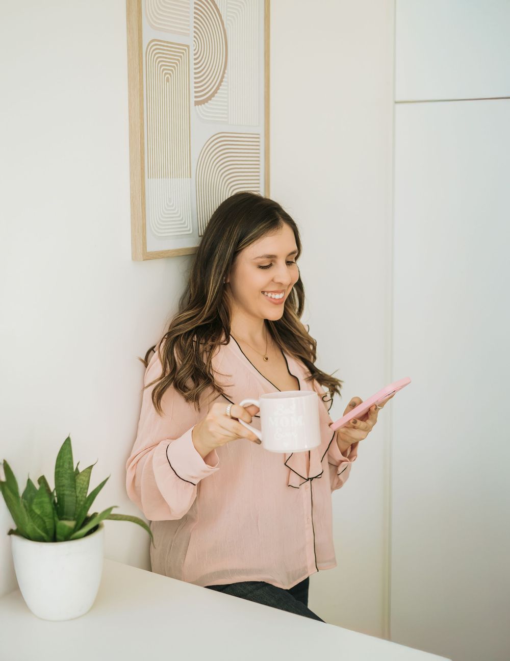 A person in a pink sweater looks at their phone while holding a white coffee cup near modern wall art and a potted plant.