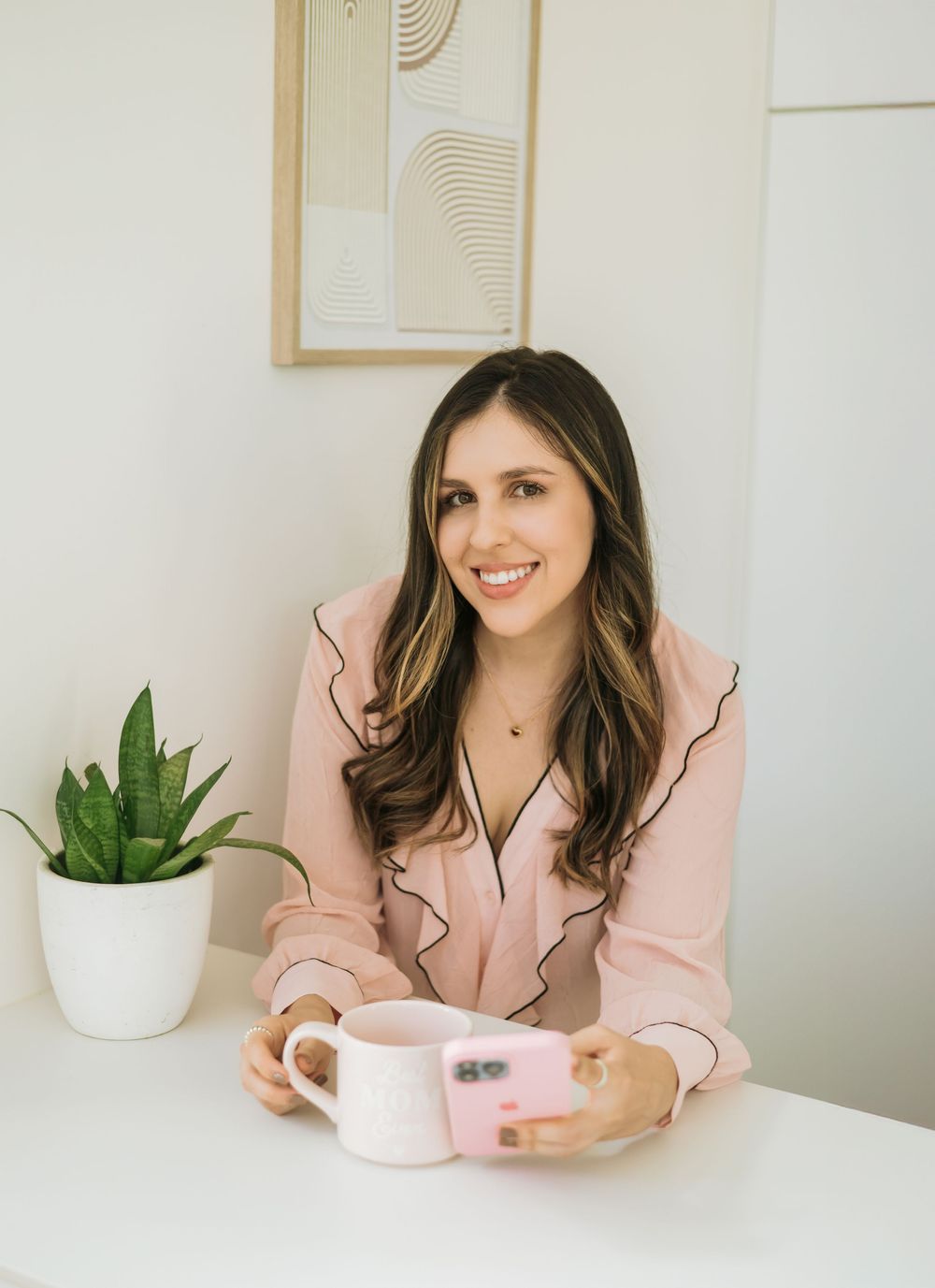 Person in pink pajama top sitting at white table with coffee mug and potted plant against minimal wall decor.