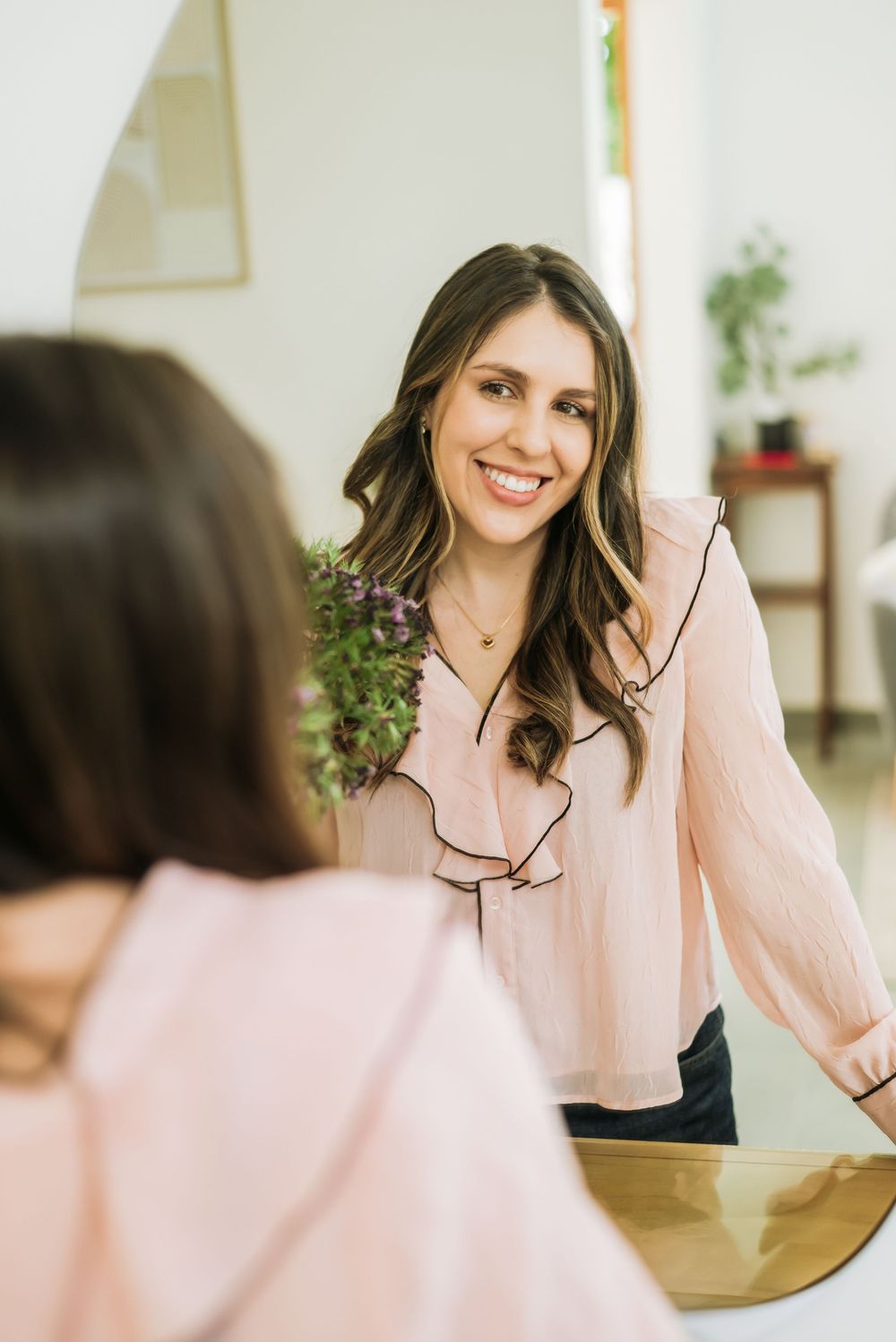 Someone wearing a pink blouse with ruffled trim looks at their reflection in a mirror while smiling in an interior setting.