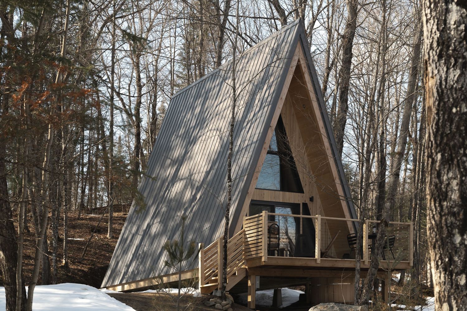 A rustic A-frame cabin with large windows and wooden deck stands among bare winter trees in a snowy forest setting.