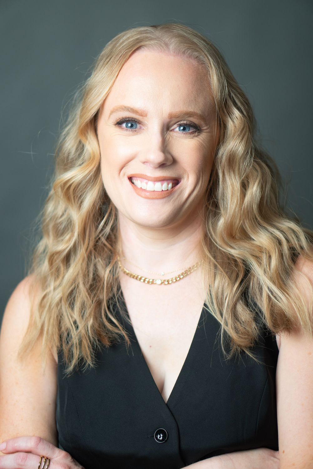 Professional headshot of a woman in a black top with wavy blonde hair and a gold necklace against a gray/blue background.