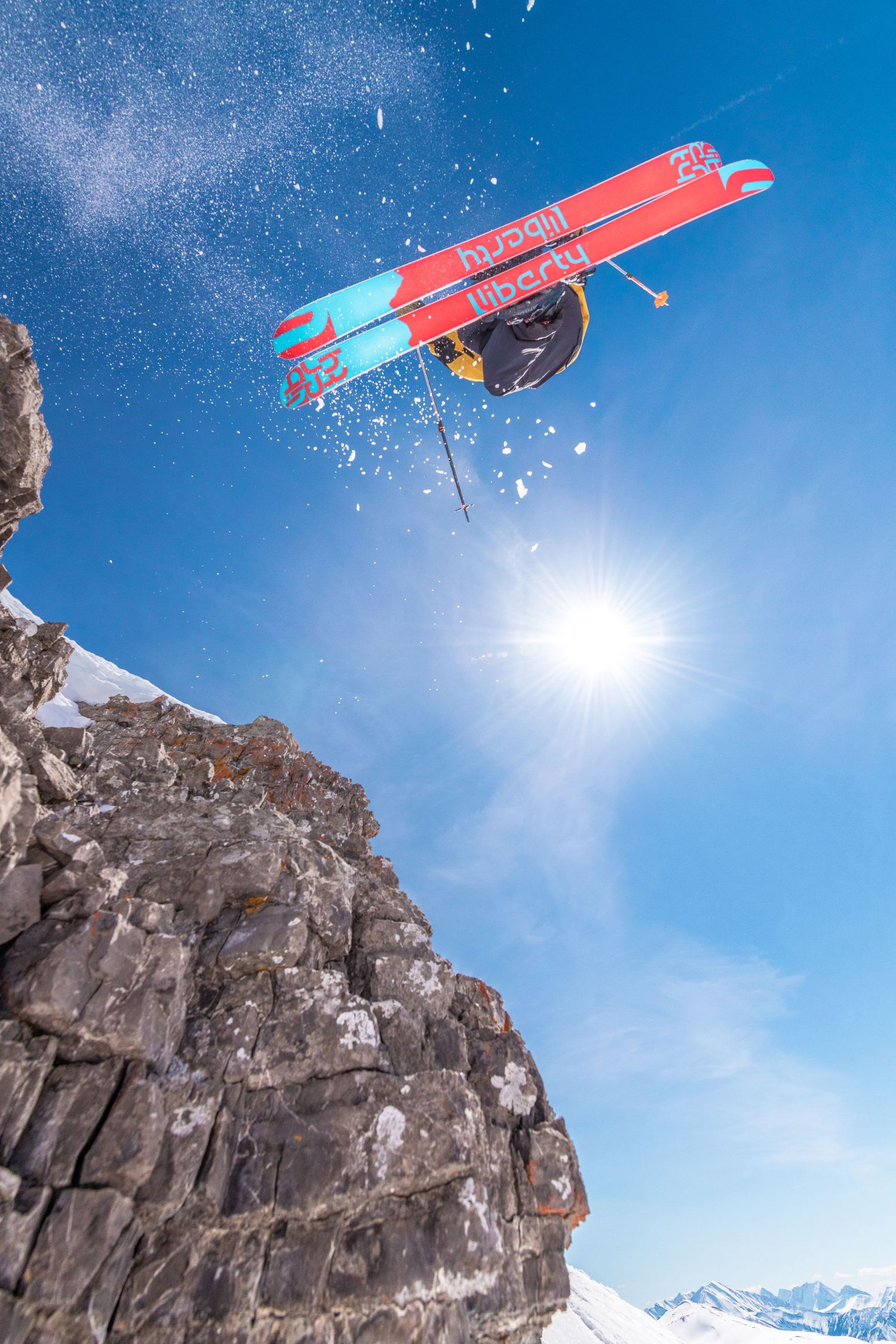 A skier performs a dramatic backflip with crossed skis against a bright blue sky and rocky mountain backdrop.
