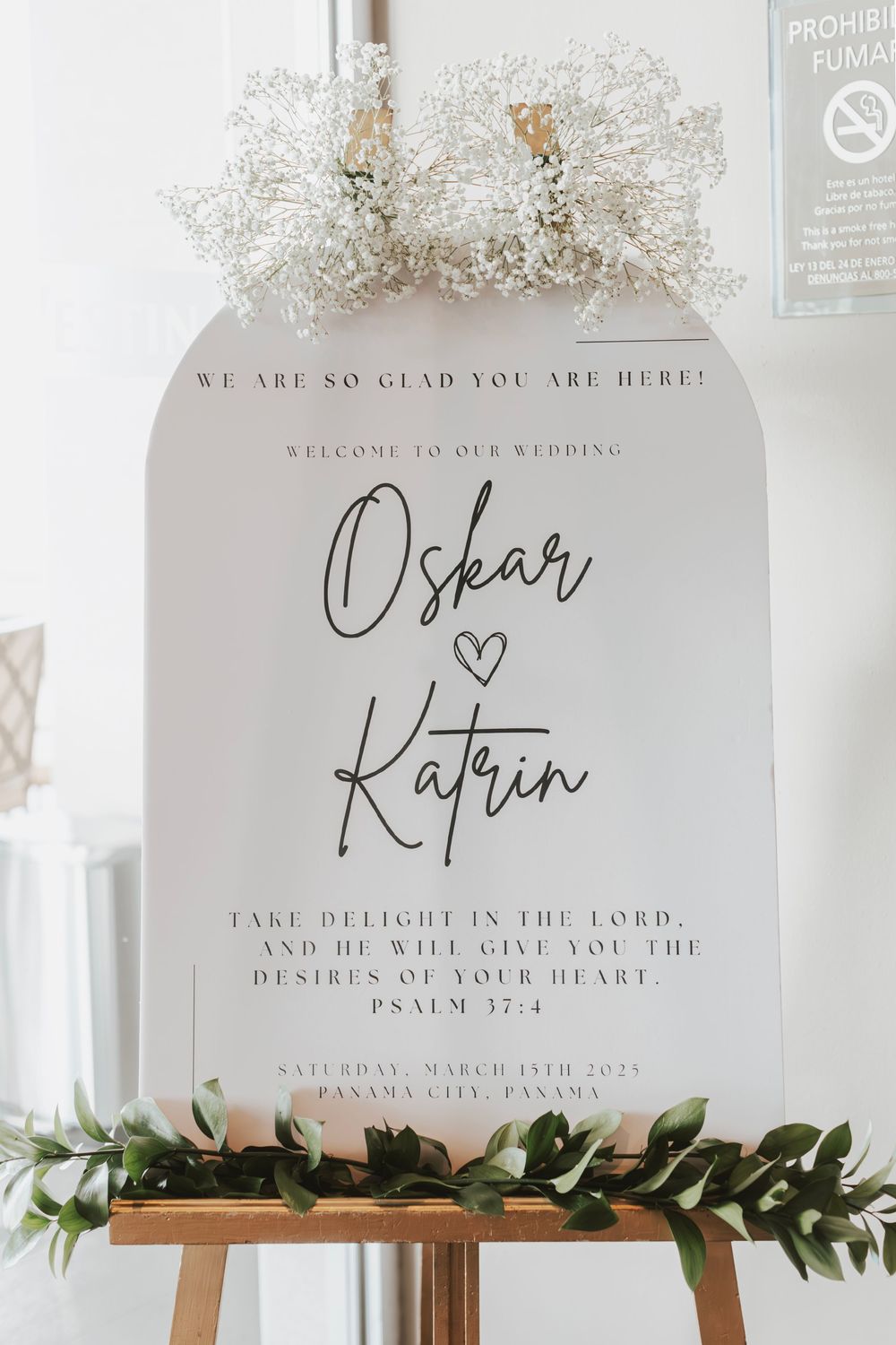 Wedding welcome sign decorated with baby's breath and greenery displaying biblical verse and couple's names.