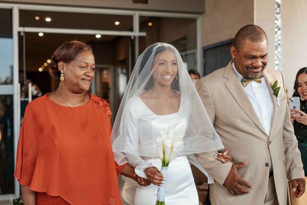 A wedding party exits a building with a bride in white dress and veil accompanied by family in orange and tan attire.