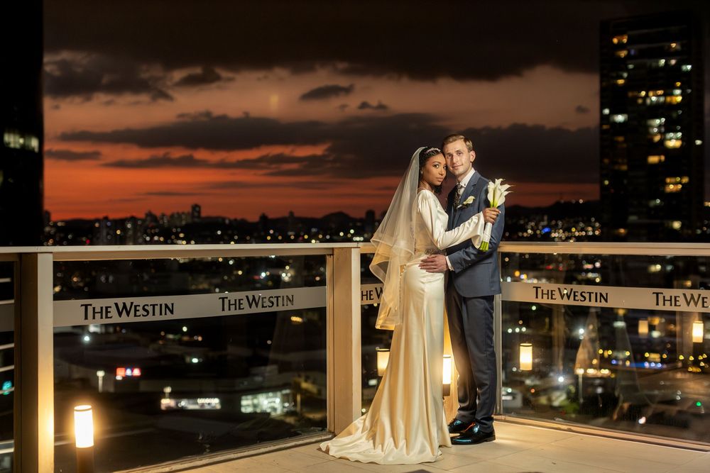 Romantic sunset wedding portrait on rooftop terrace with city lights in background.