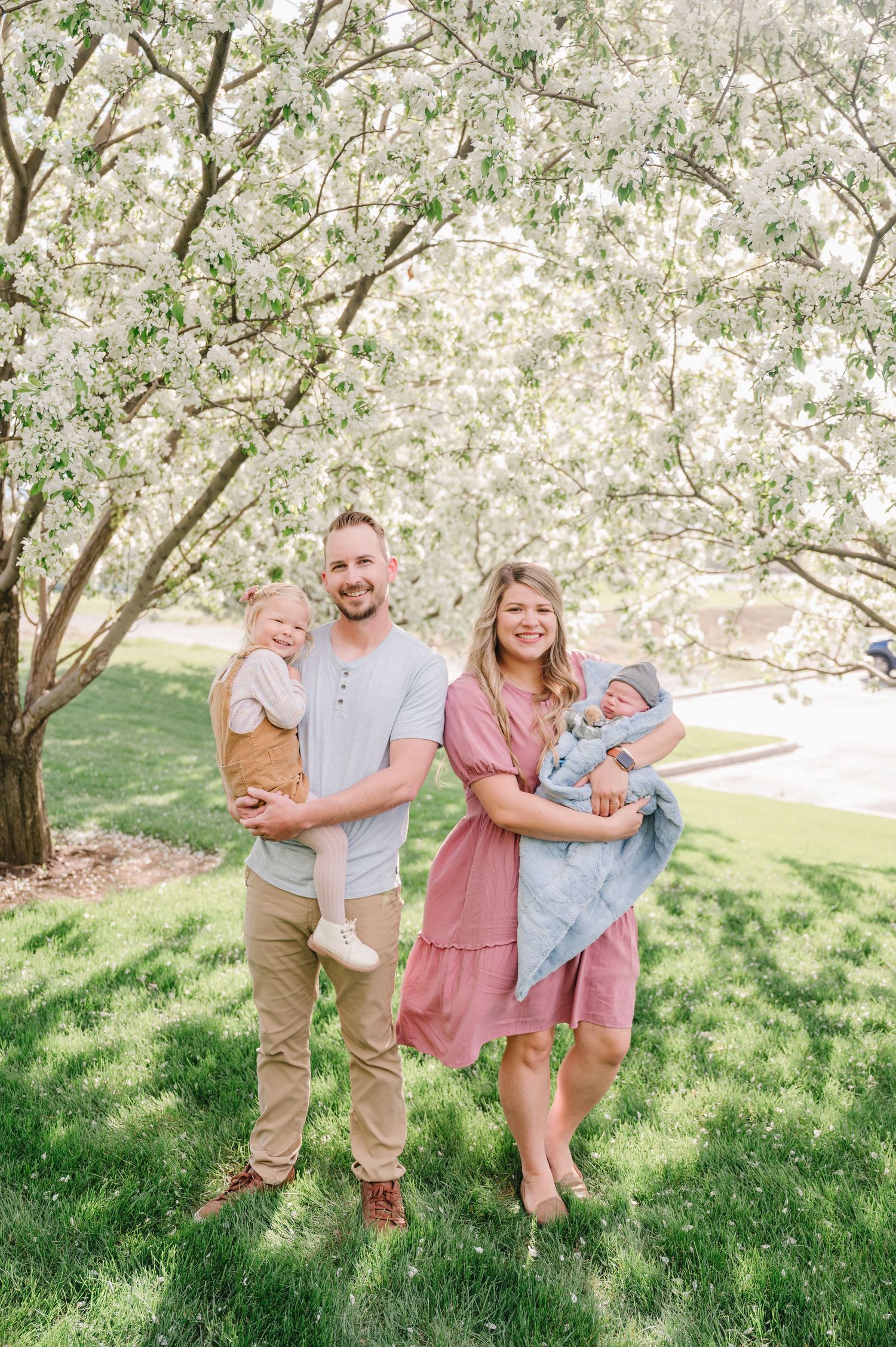 A family enjoys a spring day under blooming white trees on lush grass in a sunny outdoor setting.