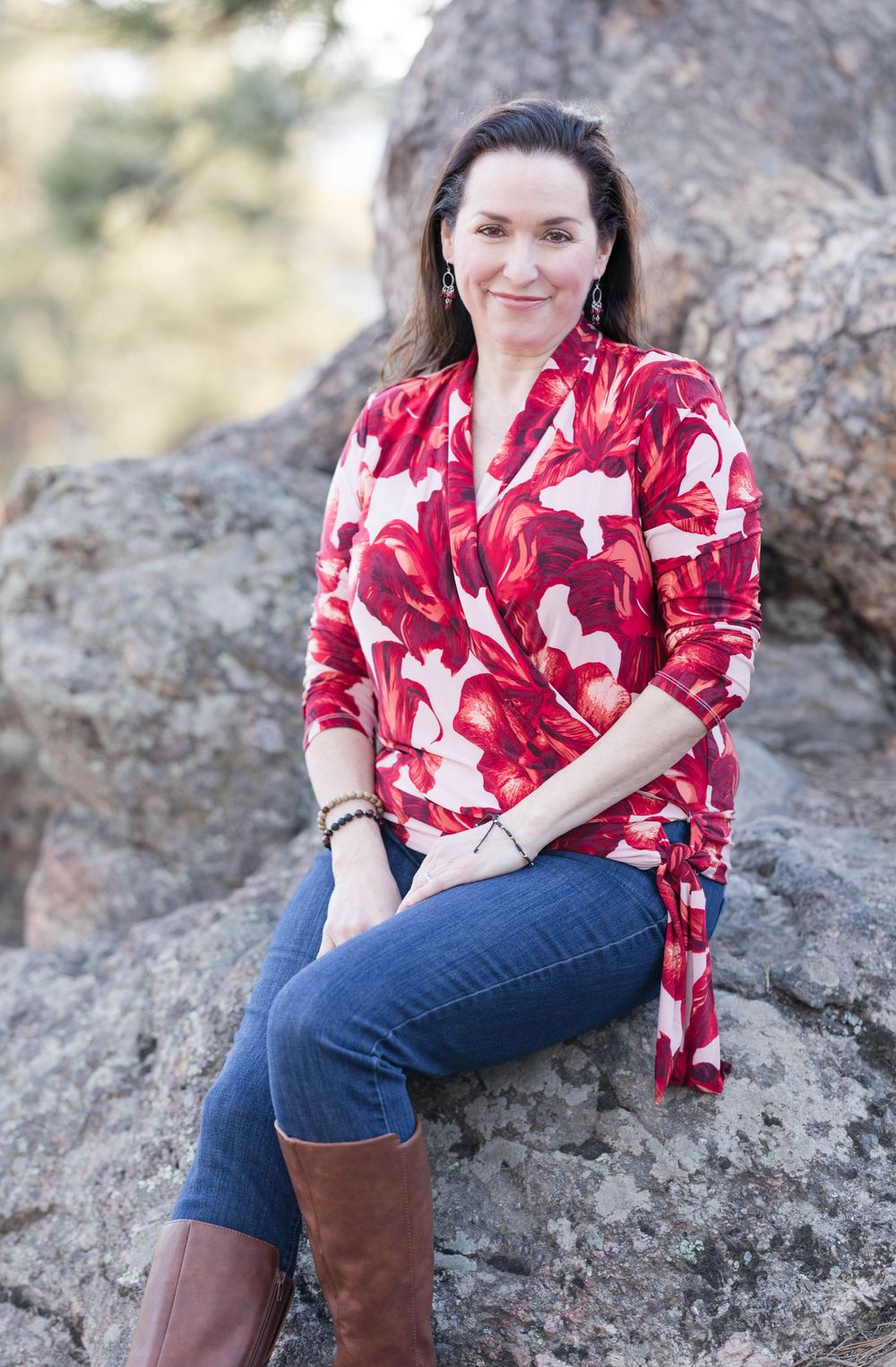 A person in a red and white patterned wrap top sits casually on a rock wearing jeans and brown boots.