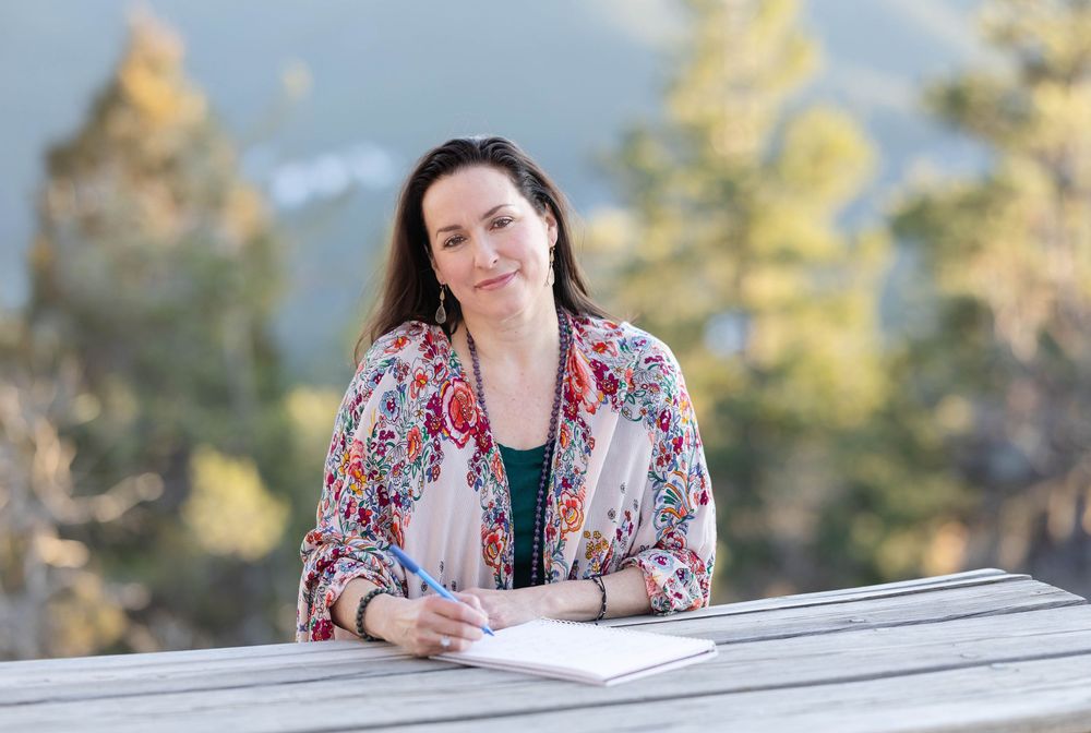 Person in floral print jacket sits at wooden table outdoors while writing with mountain backdrop and trees.