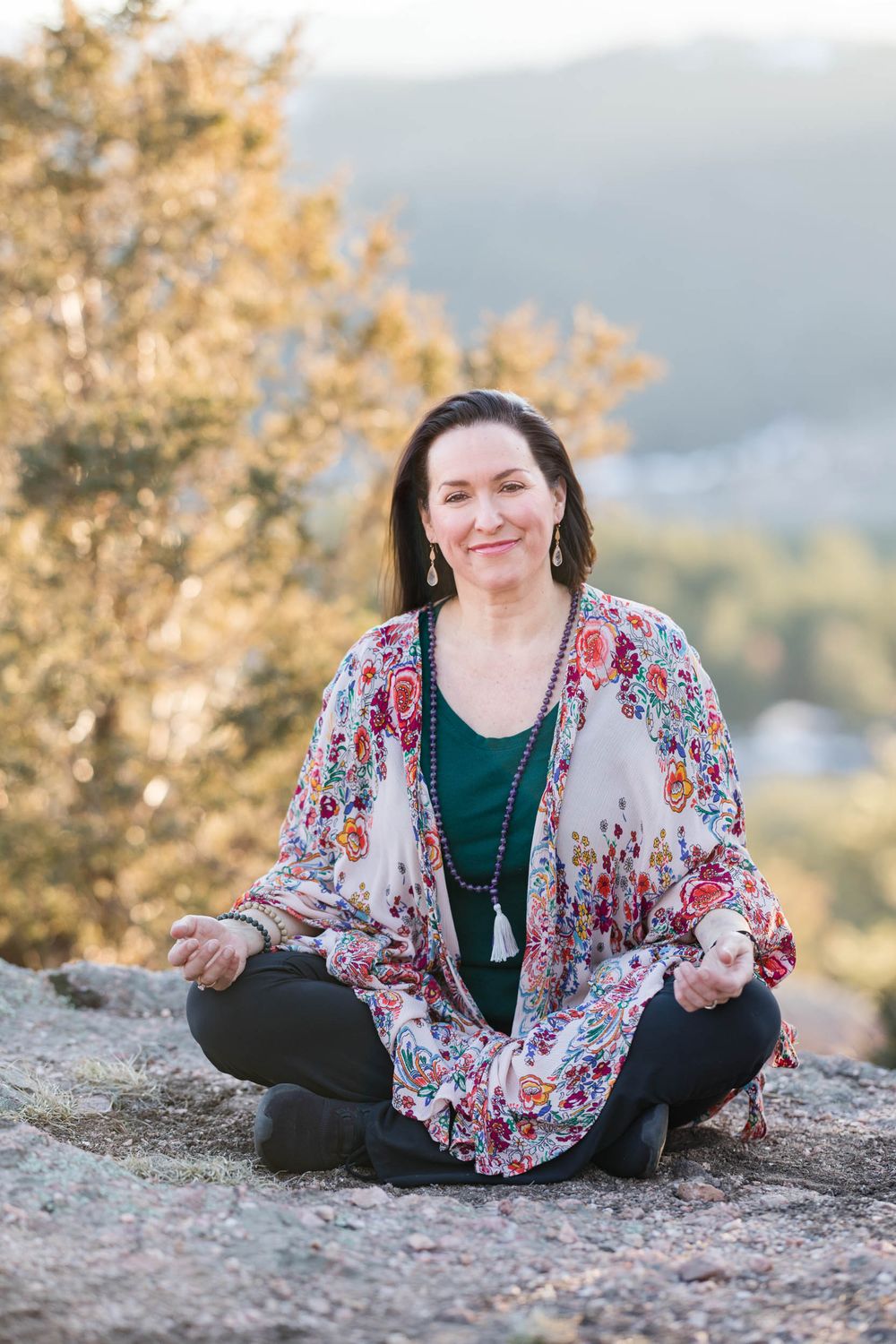 Person sits cross-legged on a rock wearing a floral kimono and green top against a nature backdrop at sunset.
