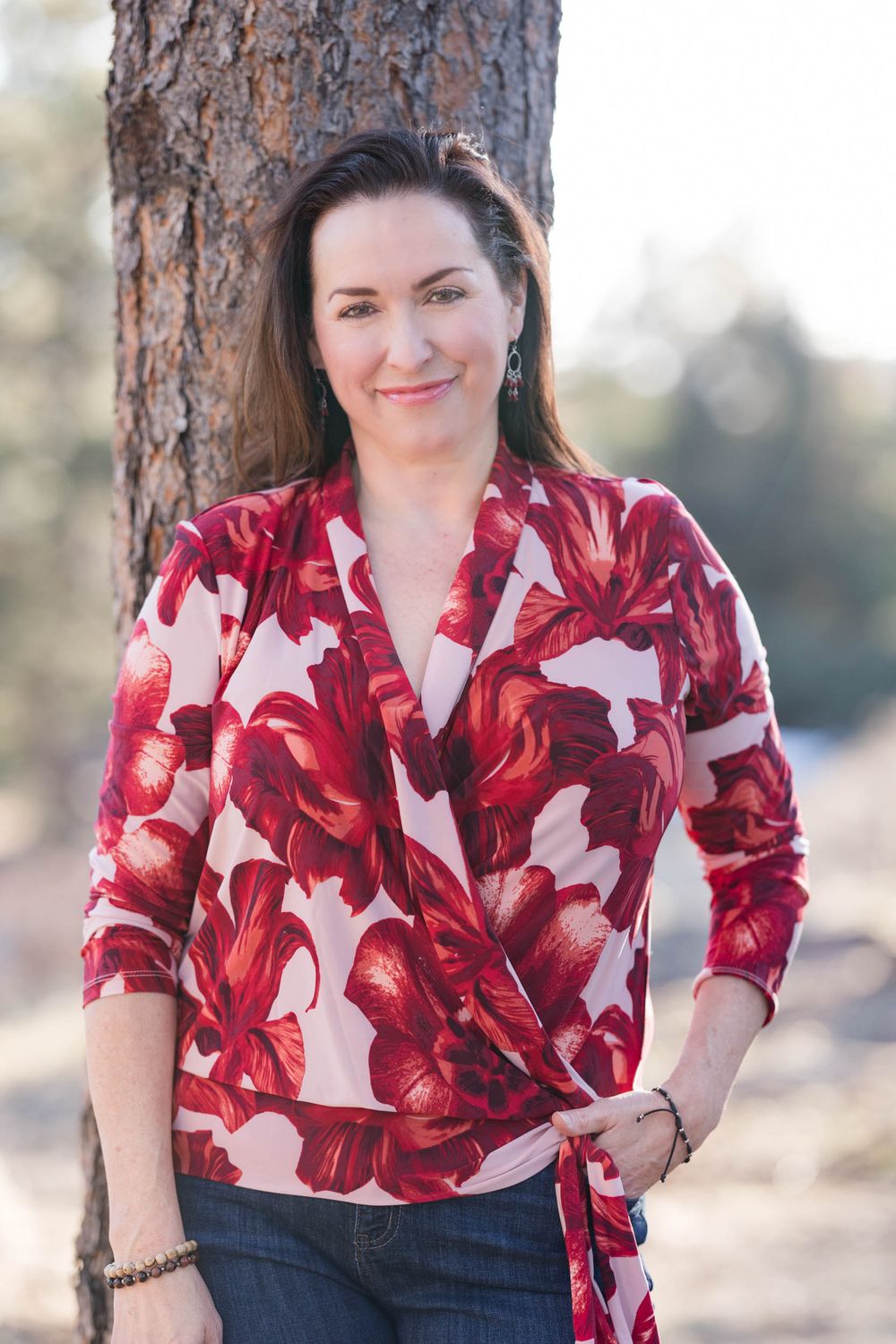 Someone poses against a tree trunk wearing a red and white floral wrap blouse paired with dark jeans in an outdoor setting.