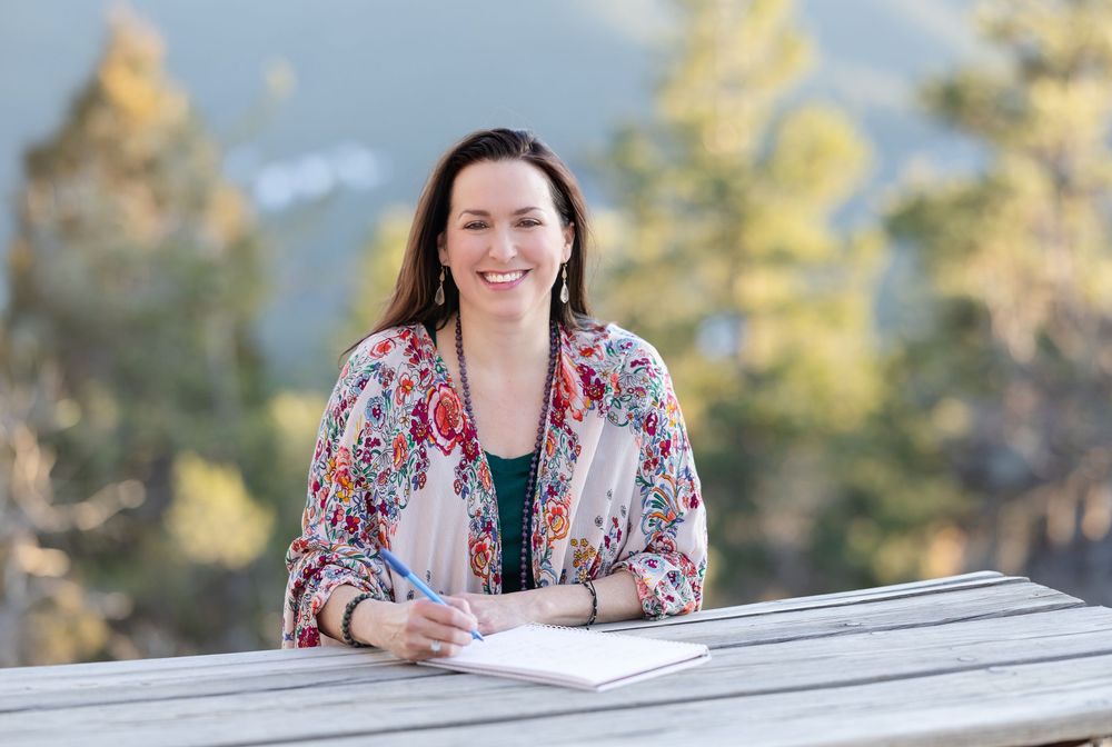 Someone wearing a colorful floral blazer sits at an outdoor table with papers on a sunny day with trees in background.