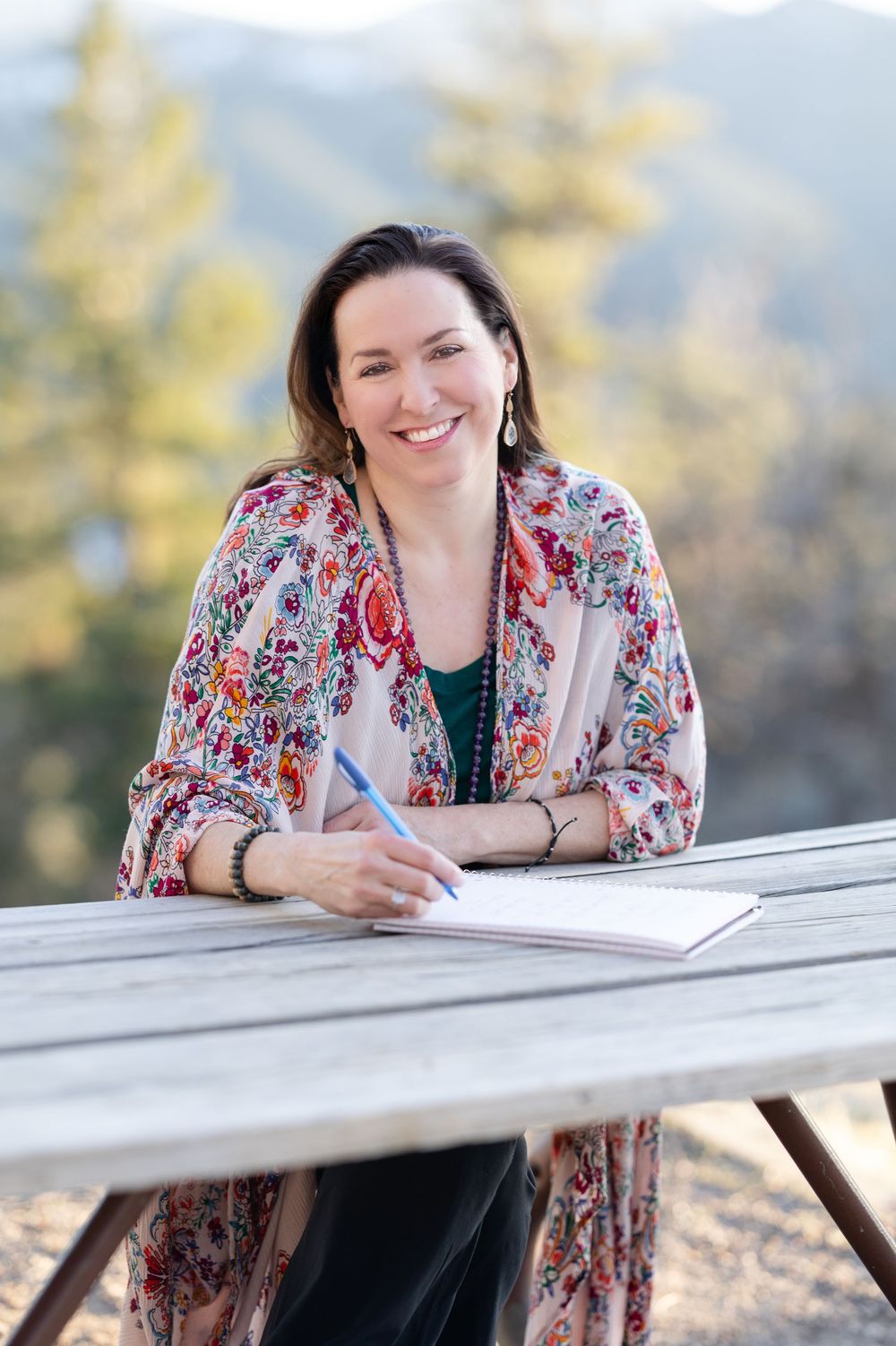 Someone writes at a wooden picnic table outdoors wearing a colorful floral kimono and green top in a mountain setting.