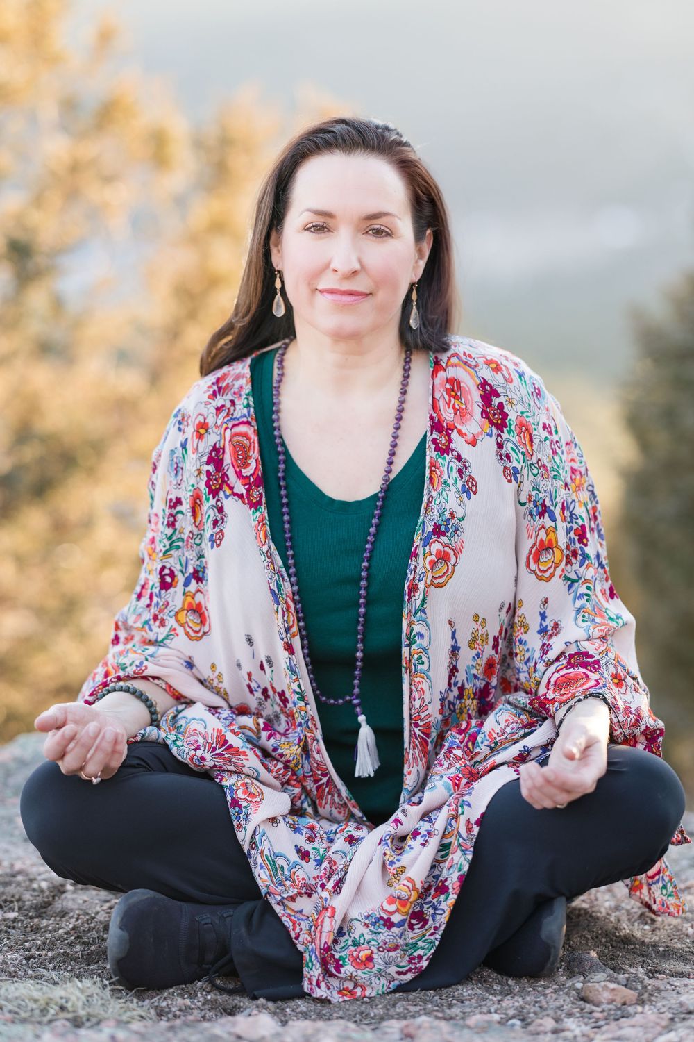 Someone sits cross-legged outdoors in meditation pose wearing a flowy floral kimono and green top against a nature backdrop.