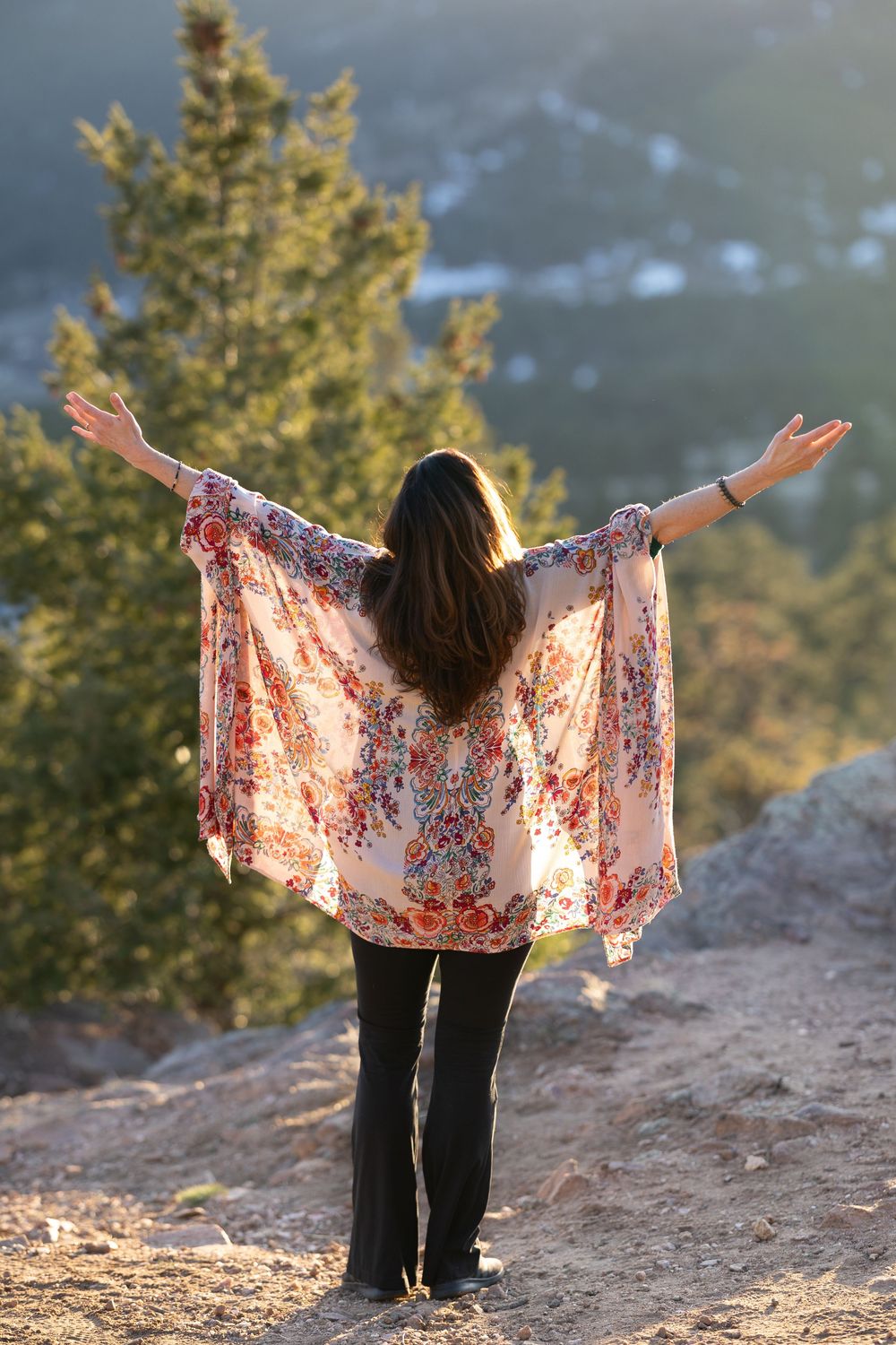 Person in floral kimono stands with outstretched arms on mountain trail during golden hour.