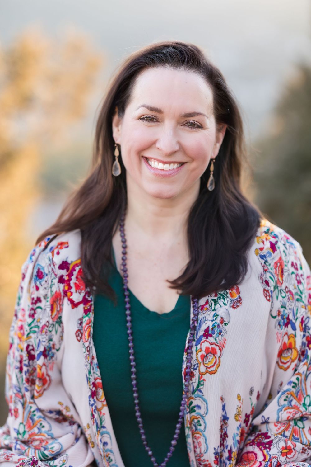 Professional headshot of someone in a floral kimono cardigan with a green top and beaded necklace against blurred background.