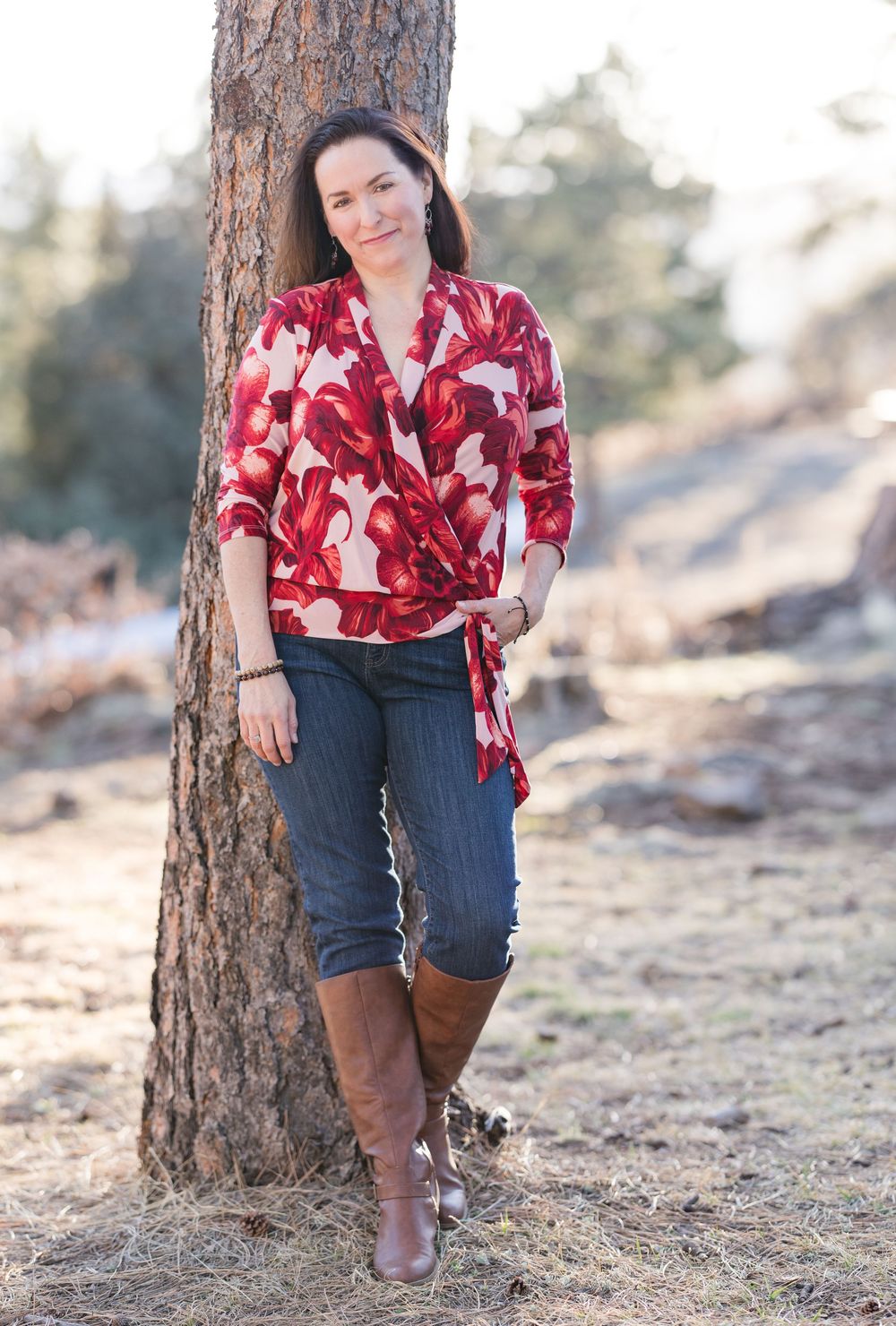 Woman in red floral wrap blouse and jeans leans against pine tree on hiking trail.