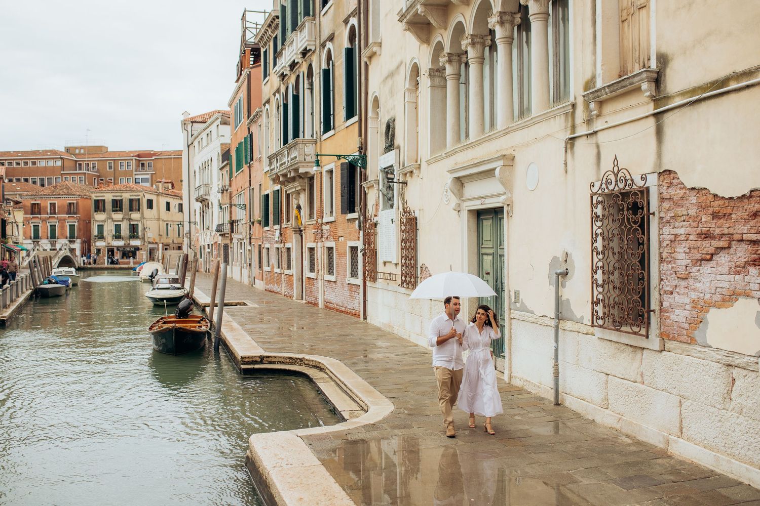 Couple in white outfits enjoying a romantic moment along a scenic Venice canal with historic buildings and gondola.