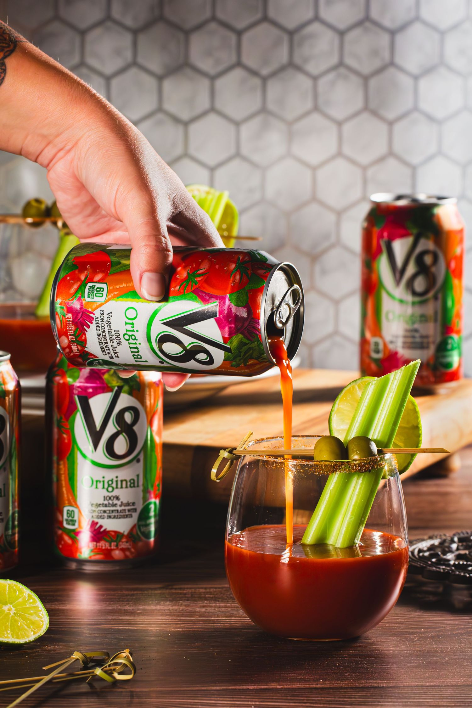 V8 vegetable juice being poured into a mocktail glass garnished with celery sticks and lime on a wooden table.