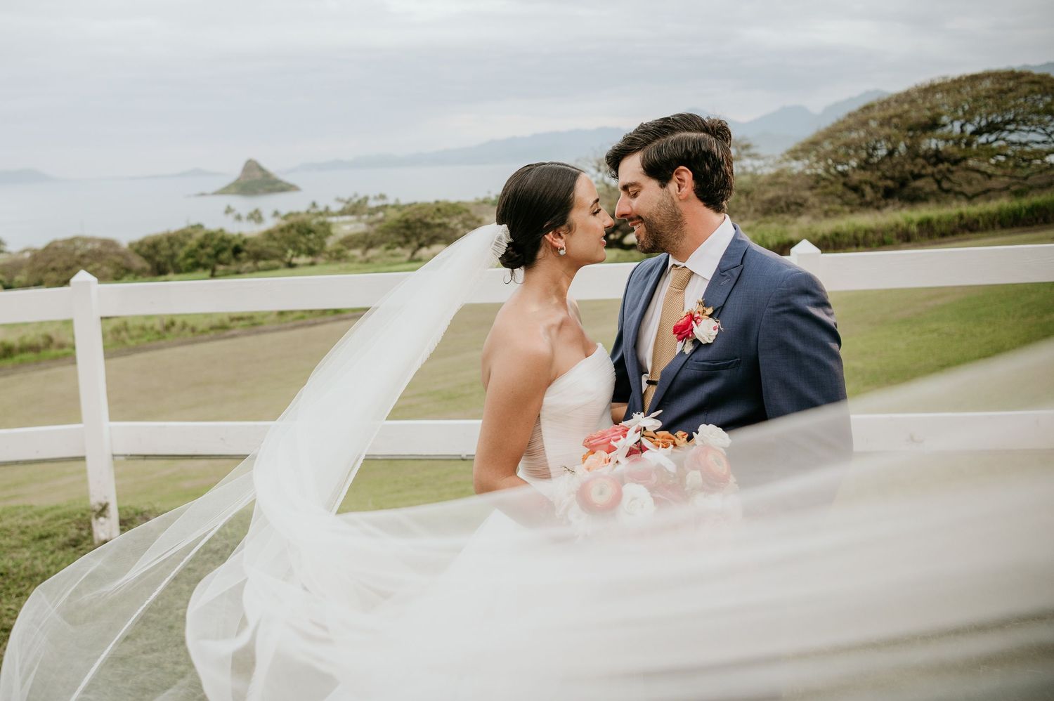 A bride and groom share an intimate moment outdoors on a cloudy day as her veil flows in the wind.