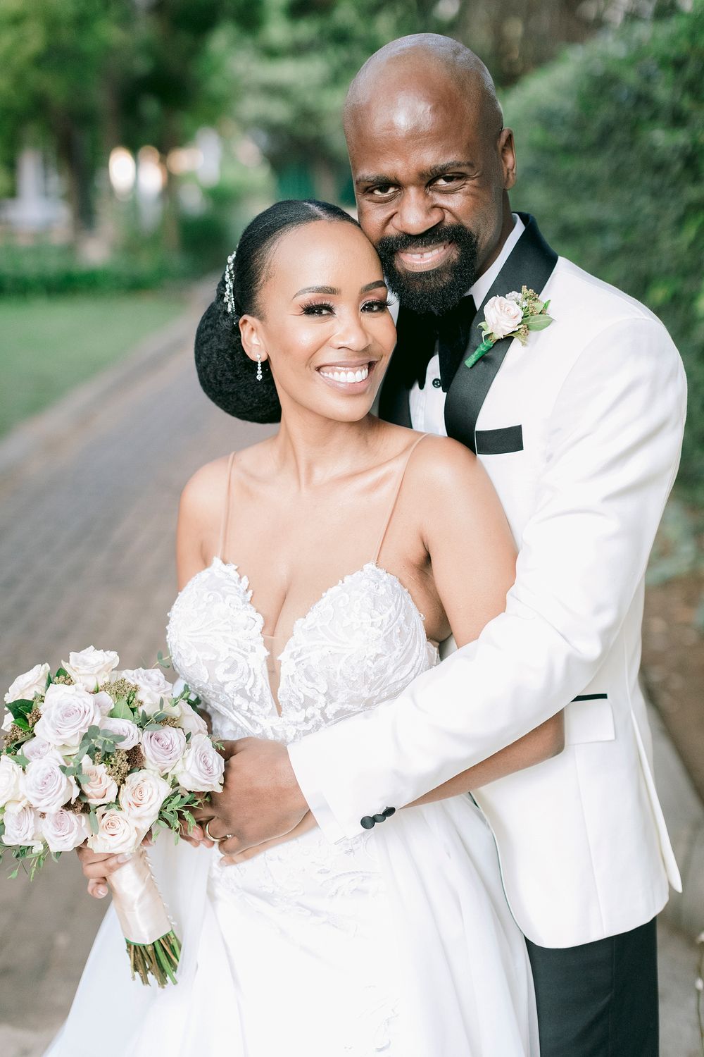Joyful bride and groom portrait outdoors, groom in white tuxedo embracing bride holding blush bouquet.