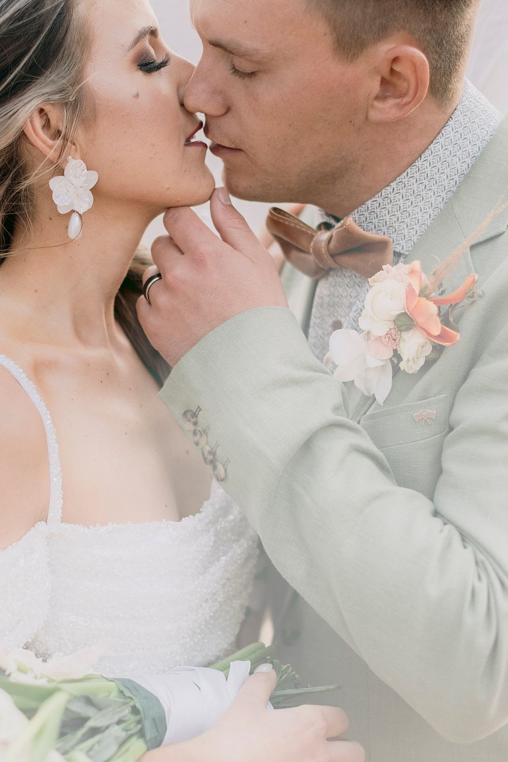 Close-up of bride and groom sharing a tender kiss, bride wearing white drop earrings and groom in a sage green suit with boutonniere.
