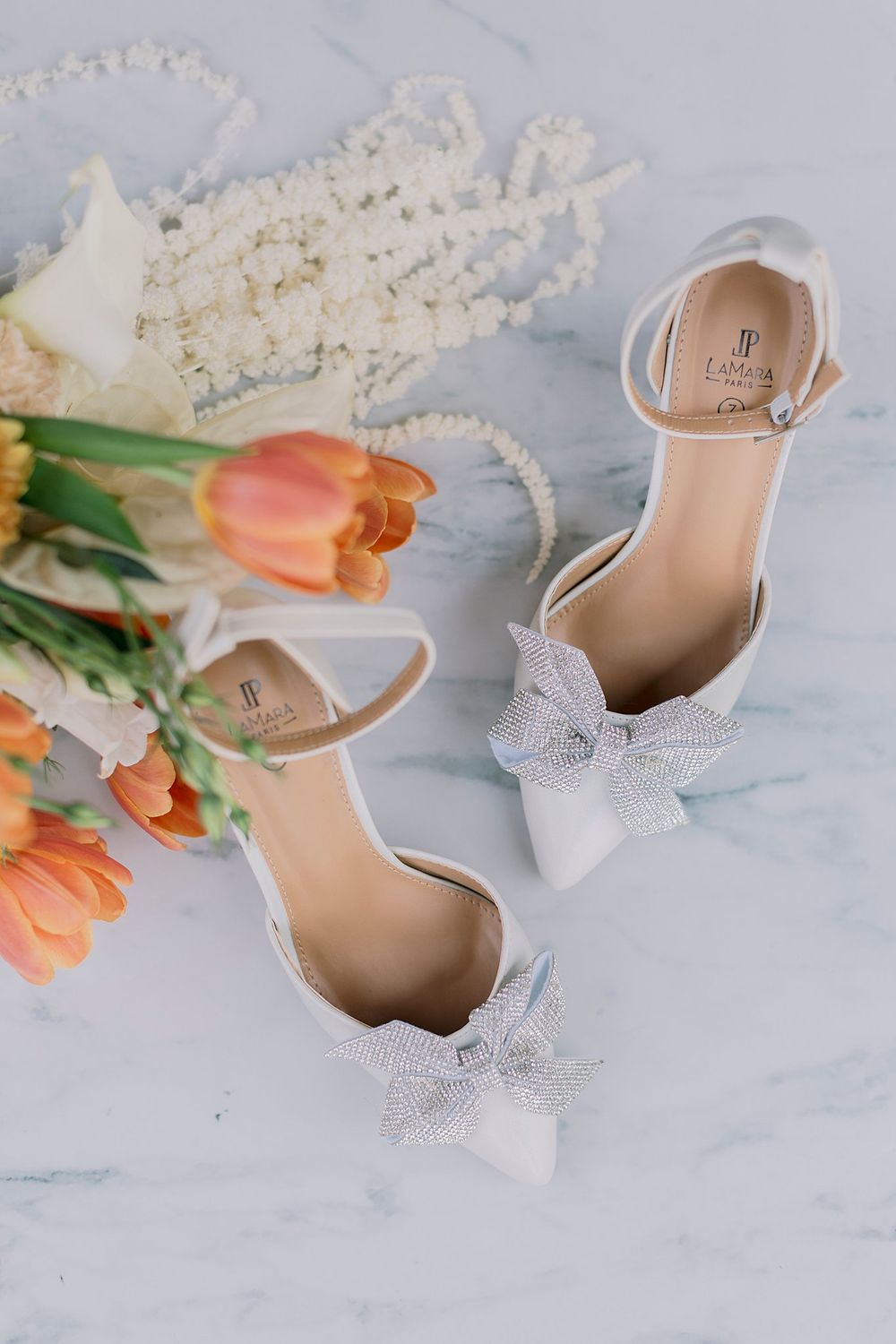 White bridal heels with crystal bows on marble surface beside orange tulips and pearl jewelry.