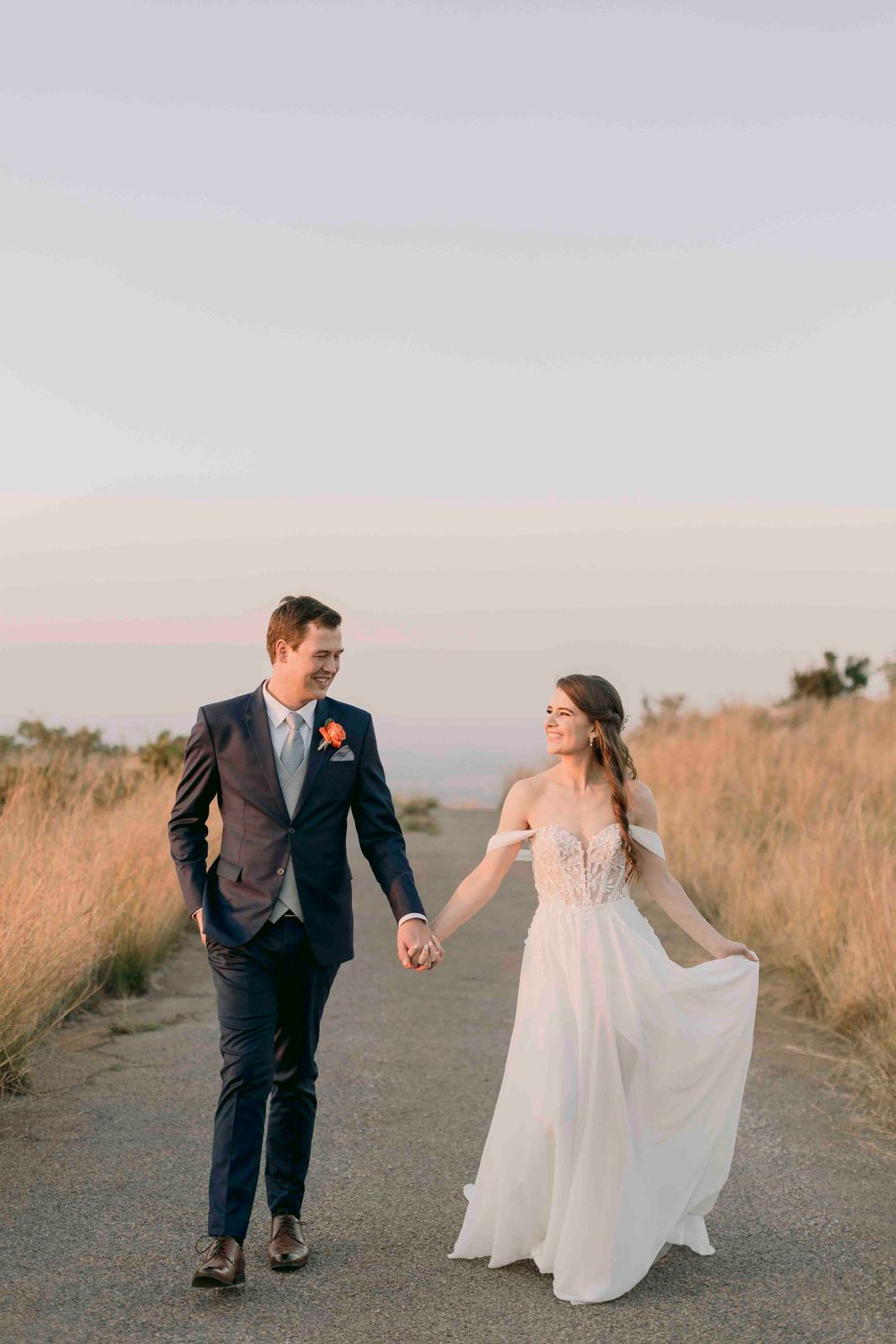 Bride and groom walking hand in hand on scenic path at dusk, exchanging loving glances.