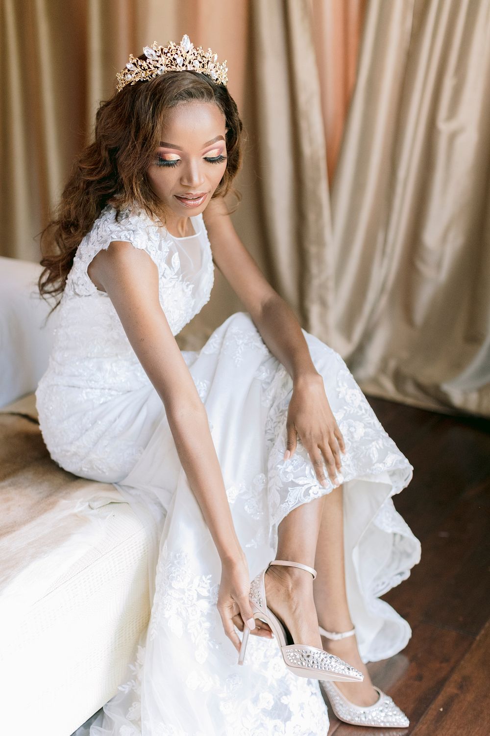 Elegant Black bride in lace wedding gown and gold tiara putting on pearl embellished bridal heels.