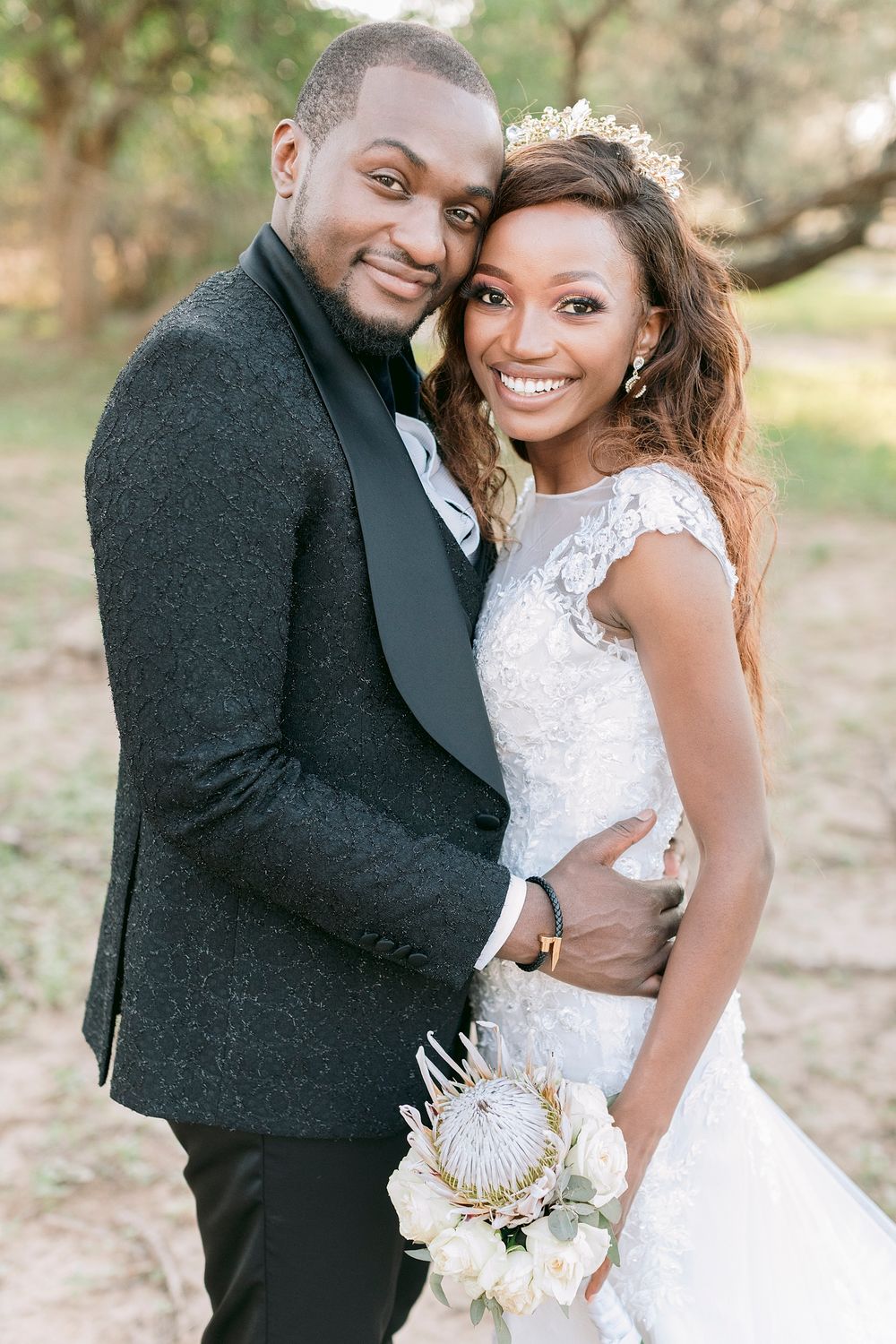 Beautiful Black bride and groom posing together outdoors, bride holding a white protea bridal bouquet.