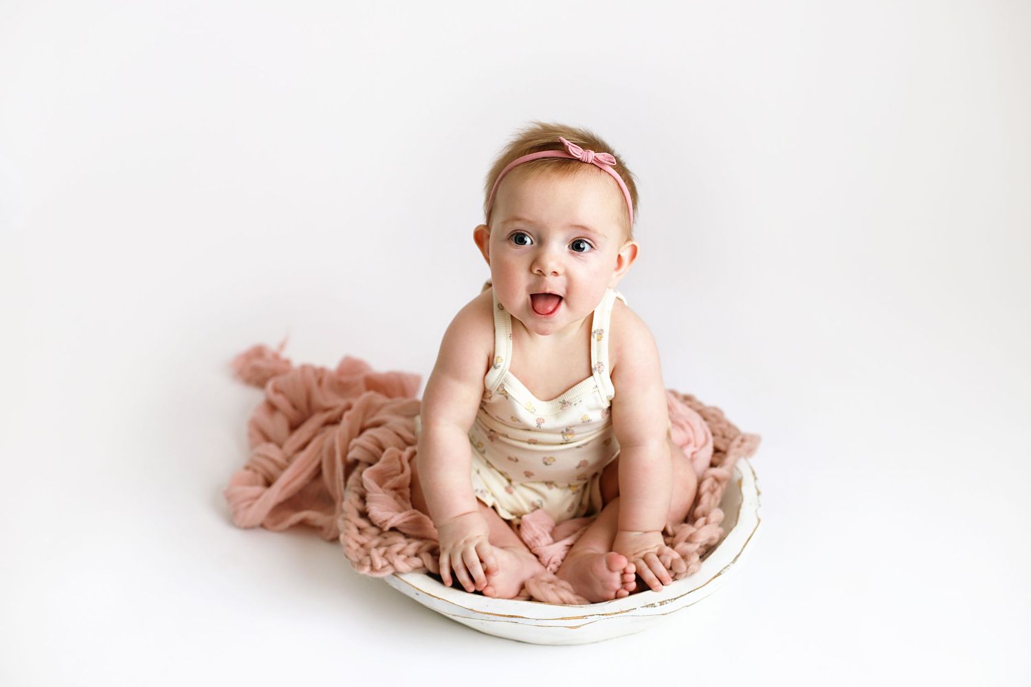 Baby in cream-colored outfit sits on white plate against pink fabric background with surprised expression.