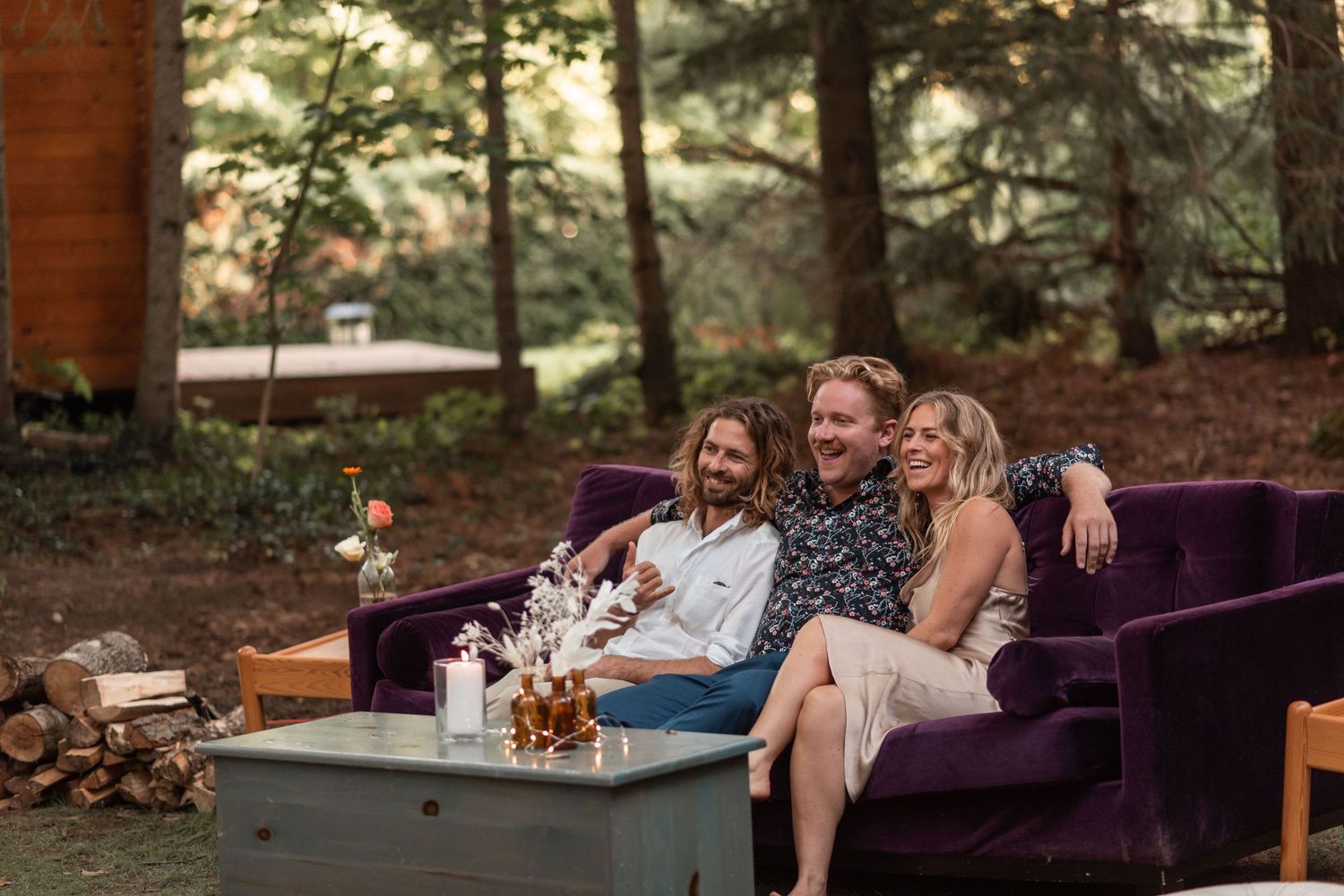 Group relaxing on plush purple outdoor sofa around coffee table with drinks at dusk in forest setting.