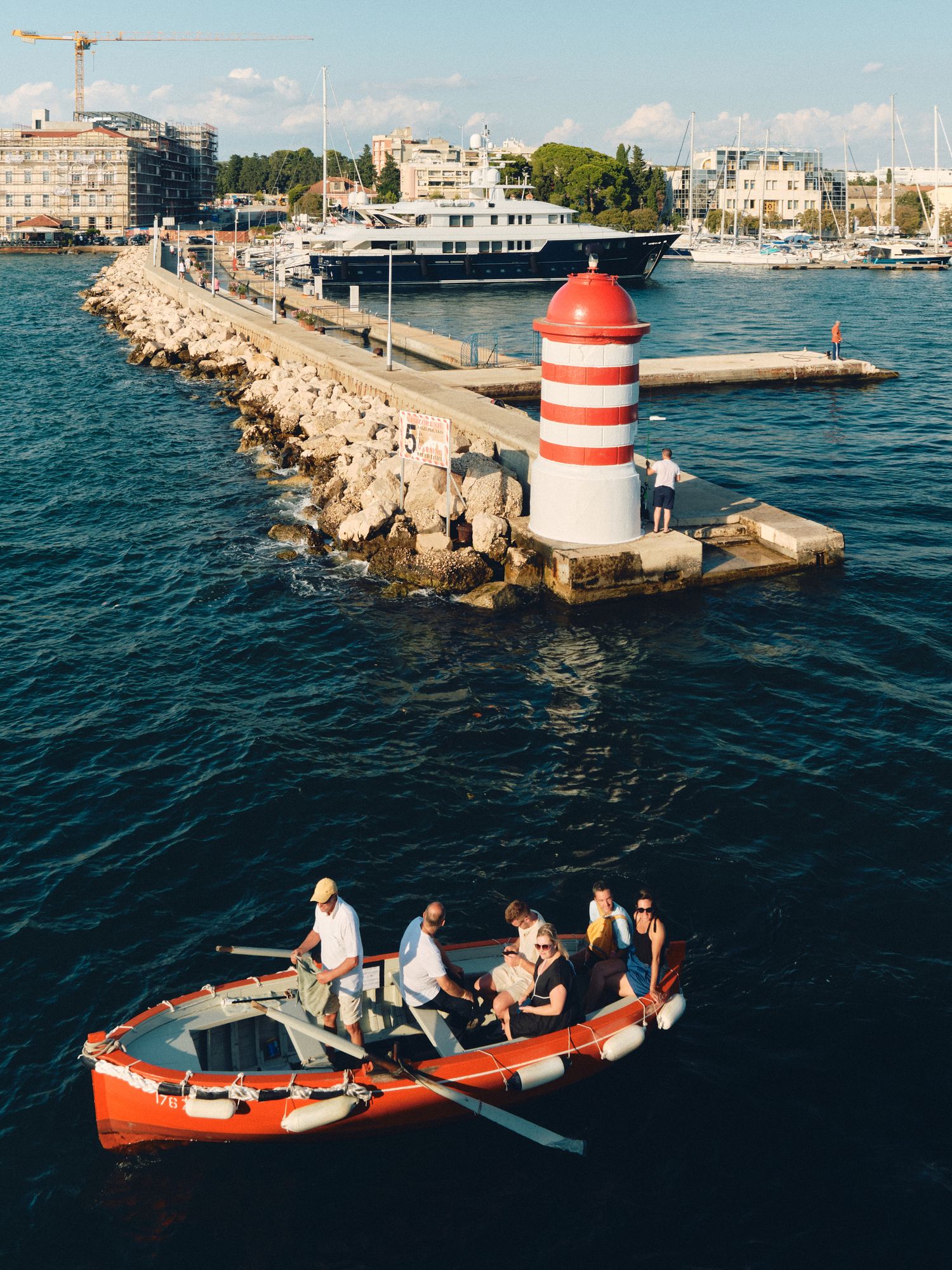 Red and white striped lighthouse at marina breakwater with luxury yacht and small boat in Mediterranean harbor.