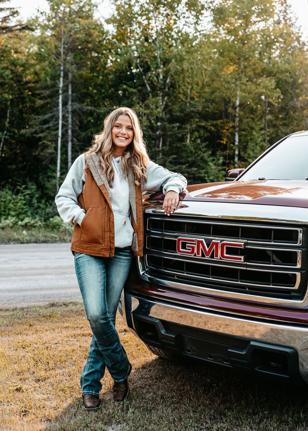 Person in brown vest and jeans posing beside front grille of black GMC truck in autumn outdoor setting.