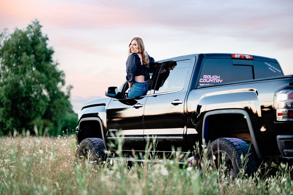 A black lifted pickup truck parked in a grassy field at sunset with trees in the background.
