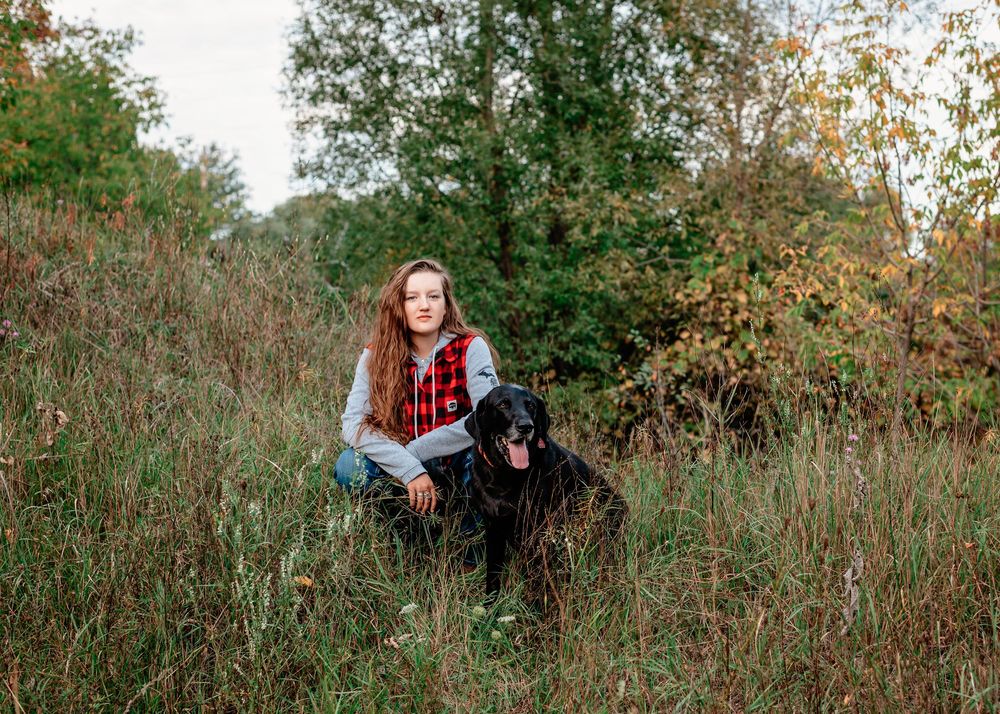 Someone sits in tall grass with a black dog on an autumn day with trees in the background.