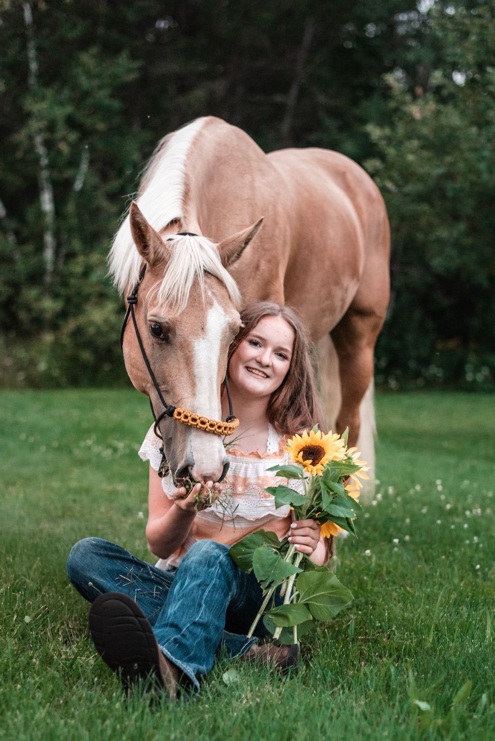 A palomino horse and someone sitting in green grass pose together with sunflowers on a summer day.