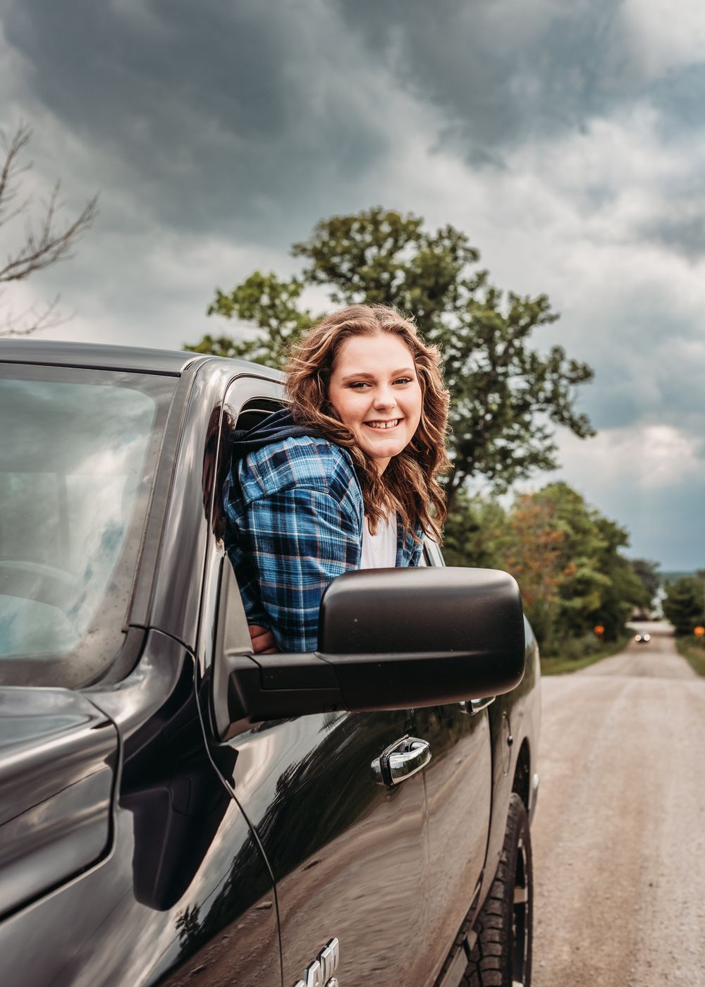 Someone in a blue plaid shirt leans out of a black truck window on a rural road under stormy clouds.