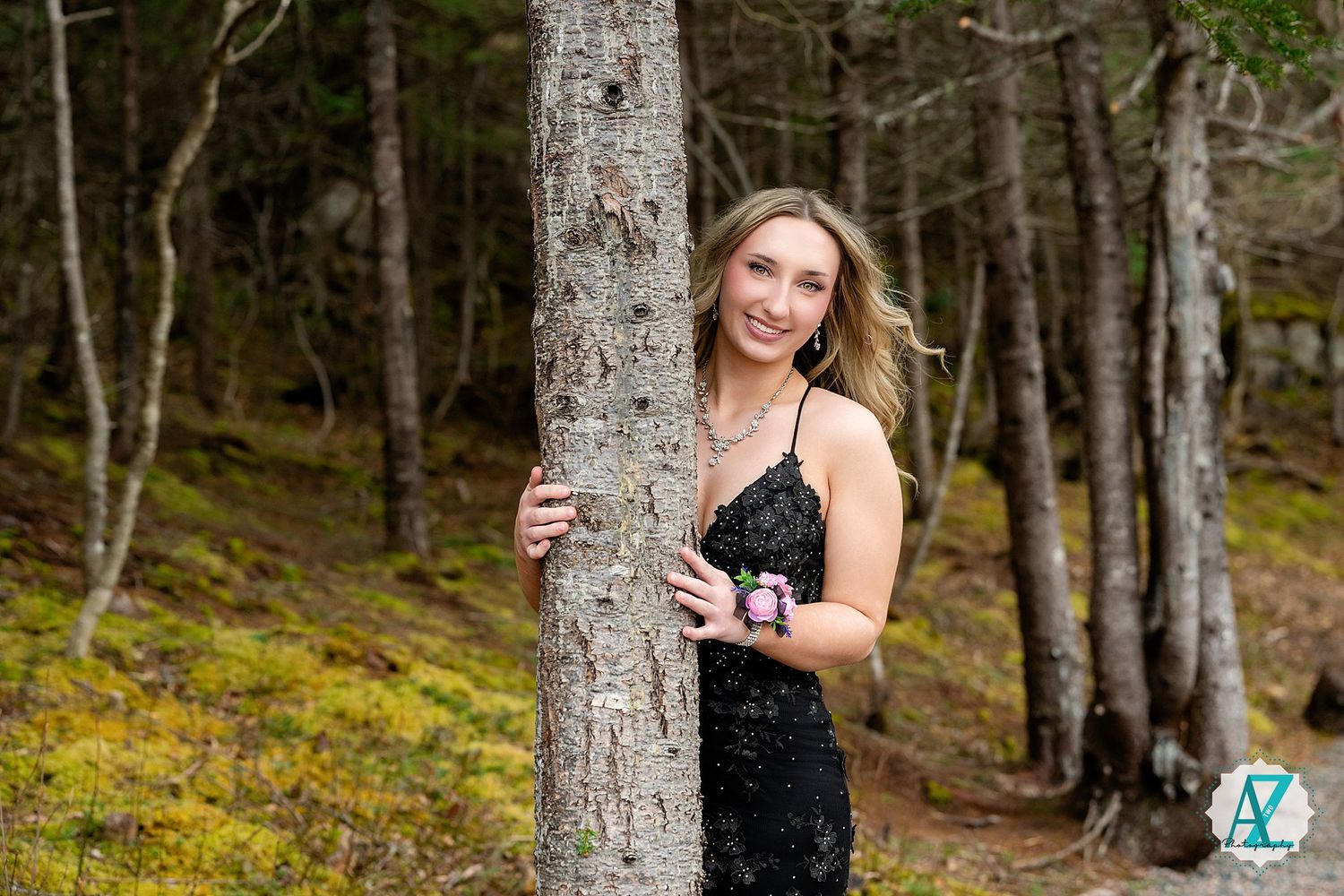 A woman in a black dress poses next to a tree trunk in a mossy forest setting during a photoshoot.