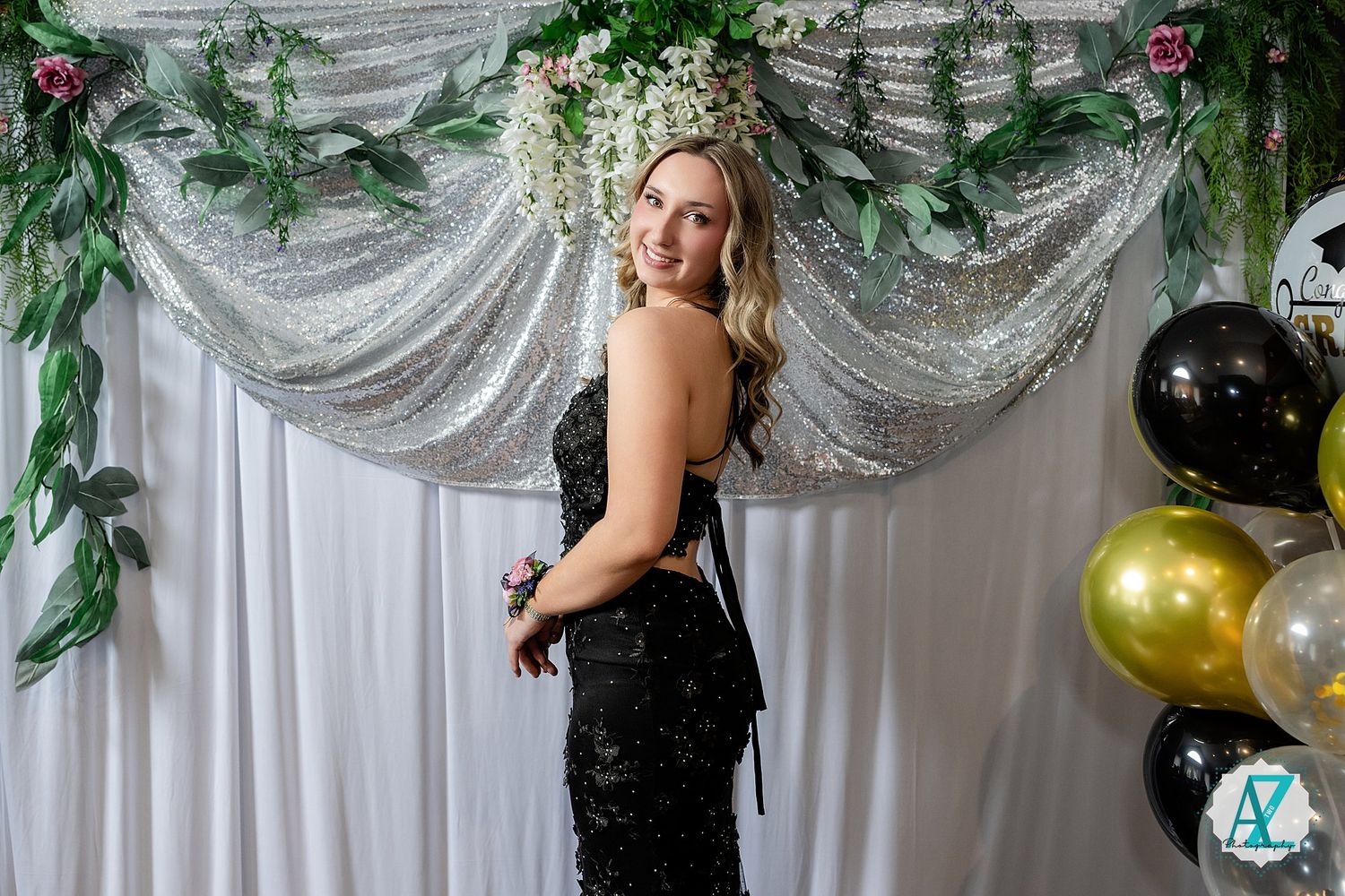 A formal event photo at a sparkly backdrop with silver draping, greenery decorations and gold balloons.