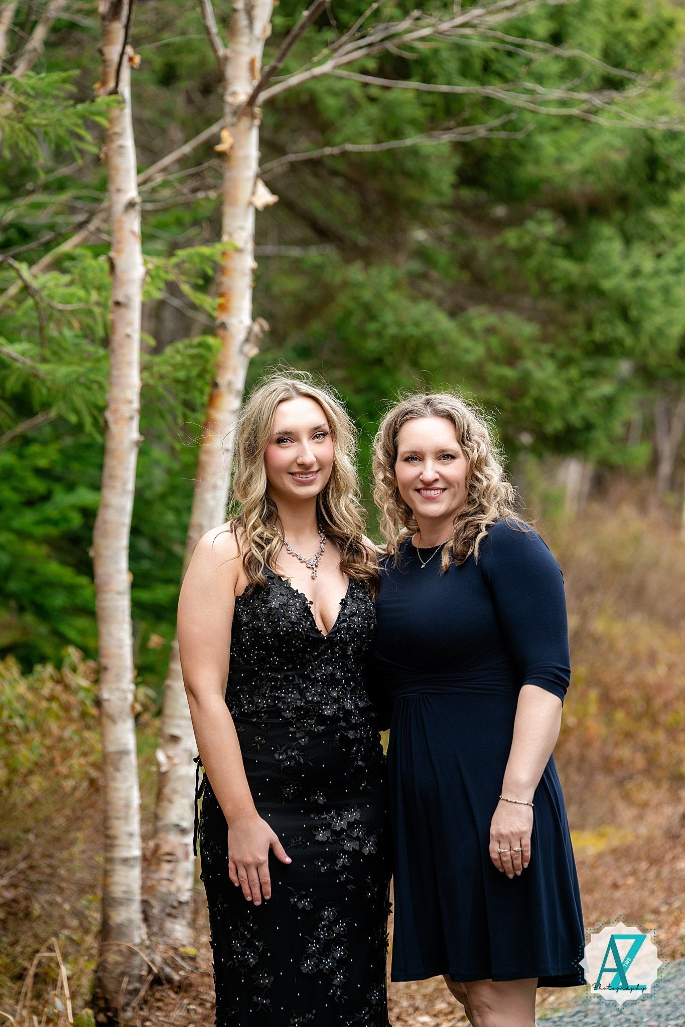 Two women in formal dresses stand together by birch trees in a wooded outdoor setting during autumn.