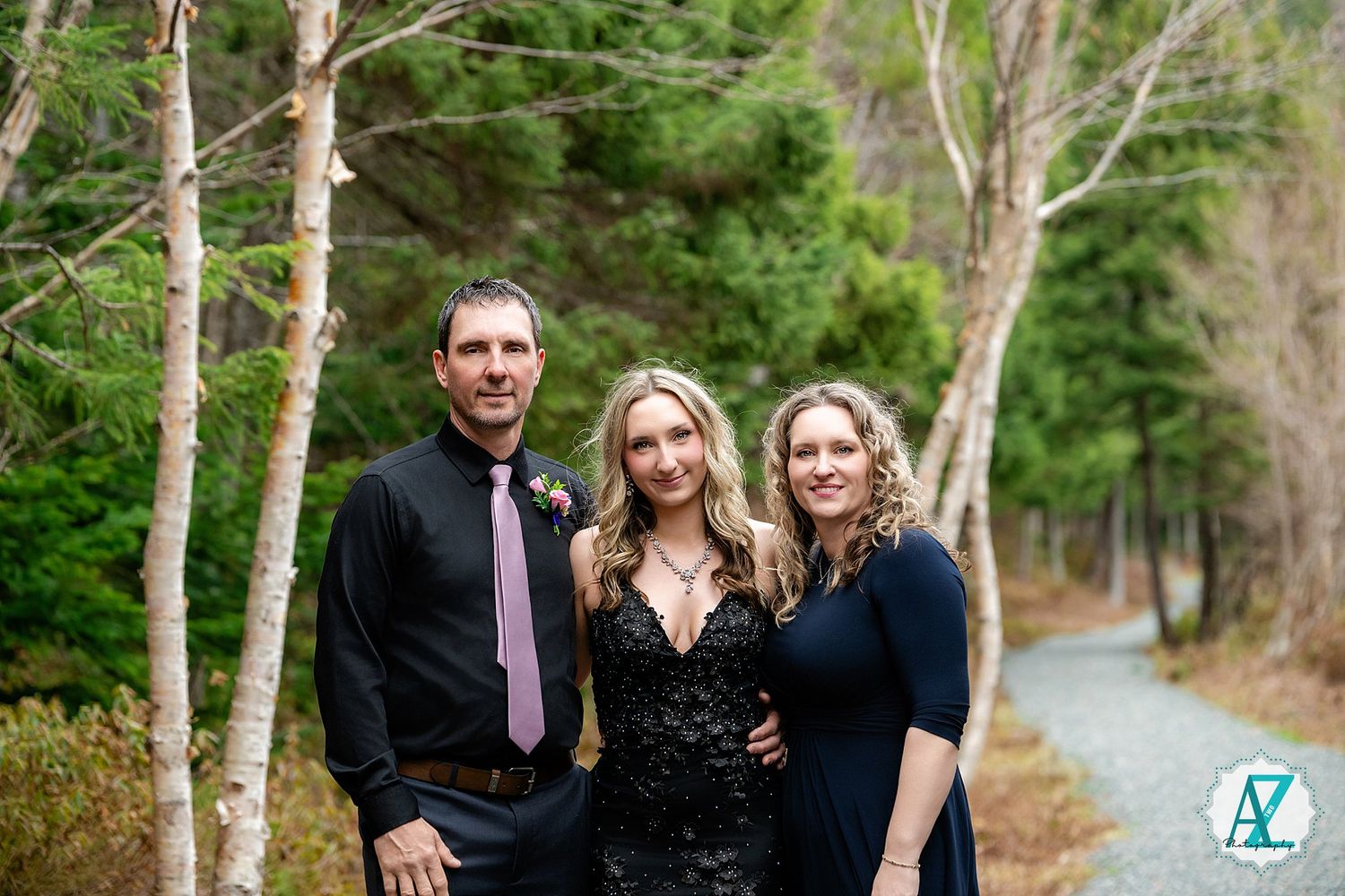 Three people in formal attire pose together on a woodland path with white birch trees and evergreens in the background.