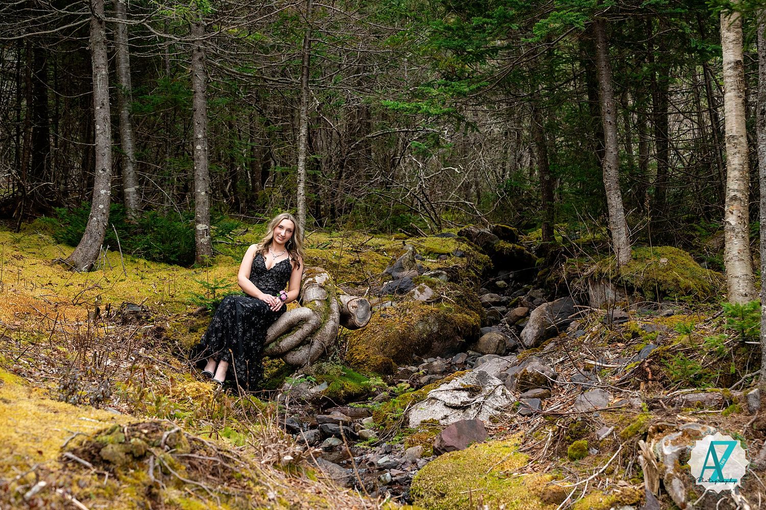 Two outdoors enthusiasts rest beside a scenic forest stream cascading over mossy rocks in a lush woodland setting.