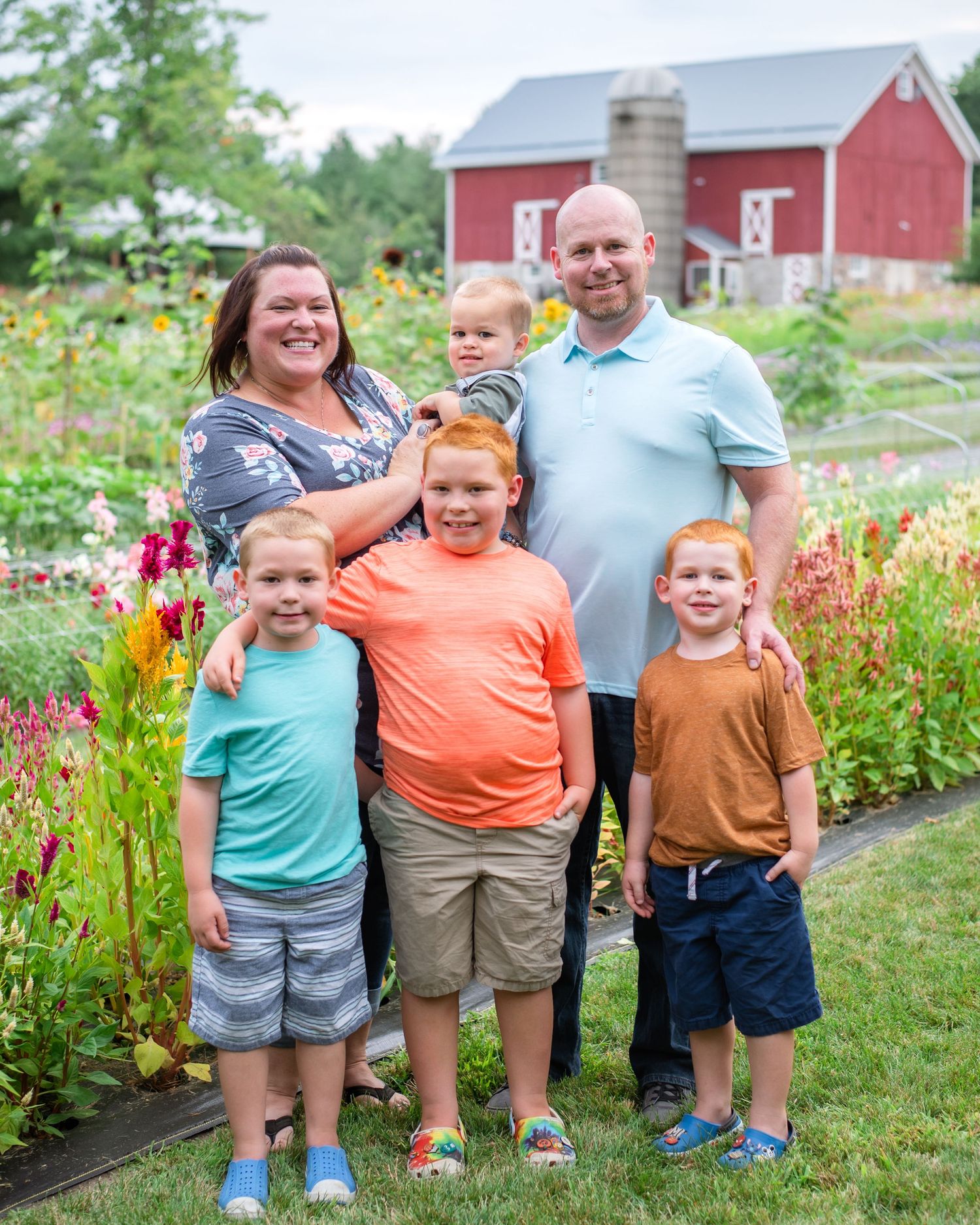 A group poses together in front of a red barn with colorful flower gardens on a sunny summer day.