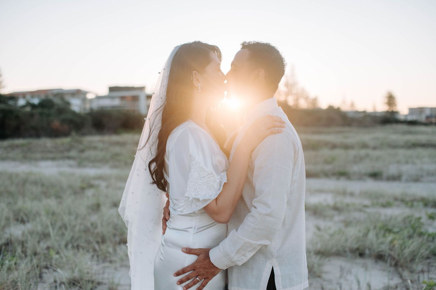 Romantic golden sunlight silhouettes couple in white outfits standing in field at sunset.