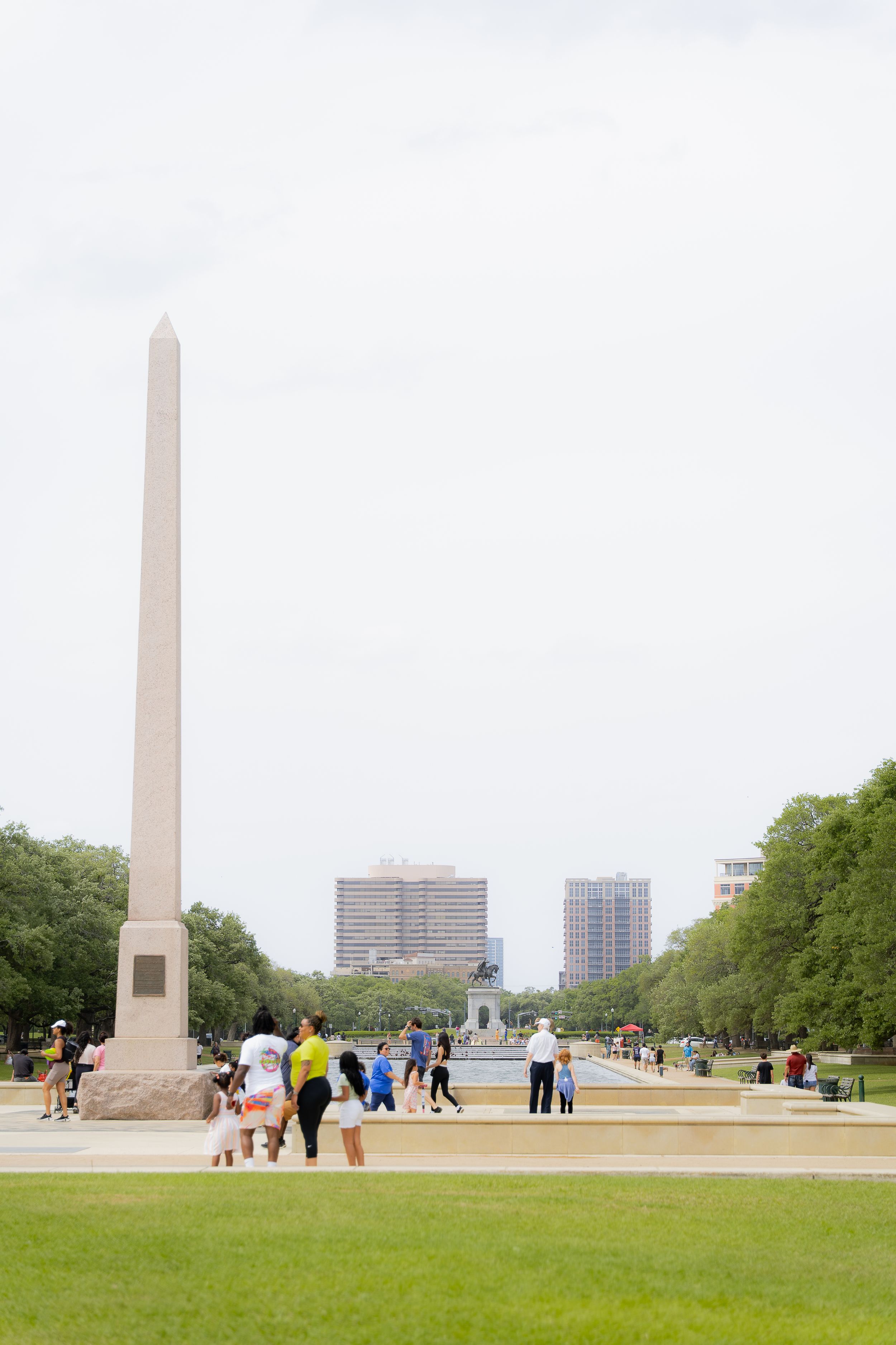 Visitors gather near a tall obelisk monument with city buildings and green trees in the background on a cloudy day.