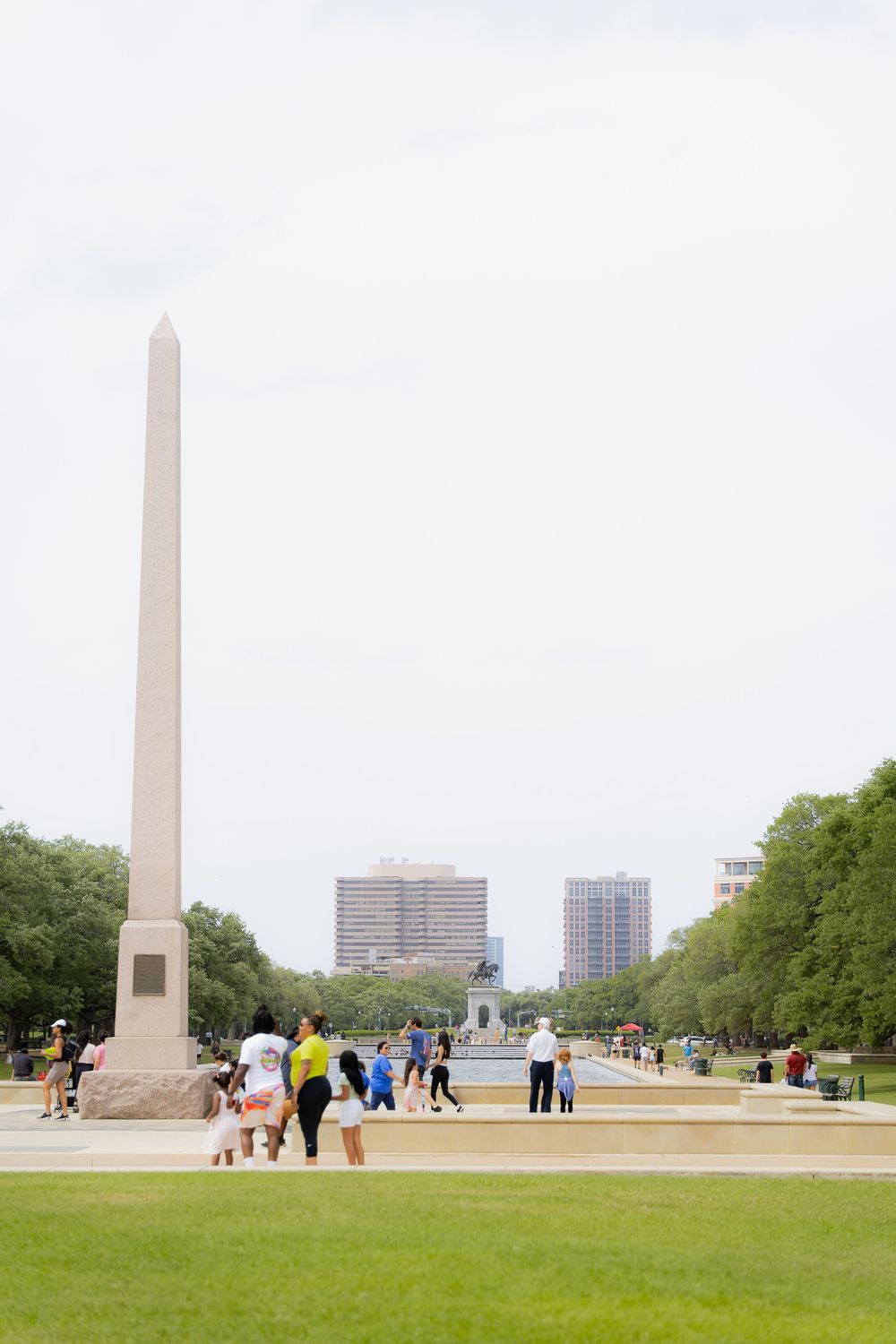 Visitors gather near a tall obelisk monument with city buildings and green trees in the background on a cloudy day.