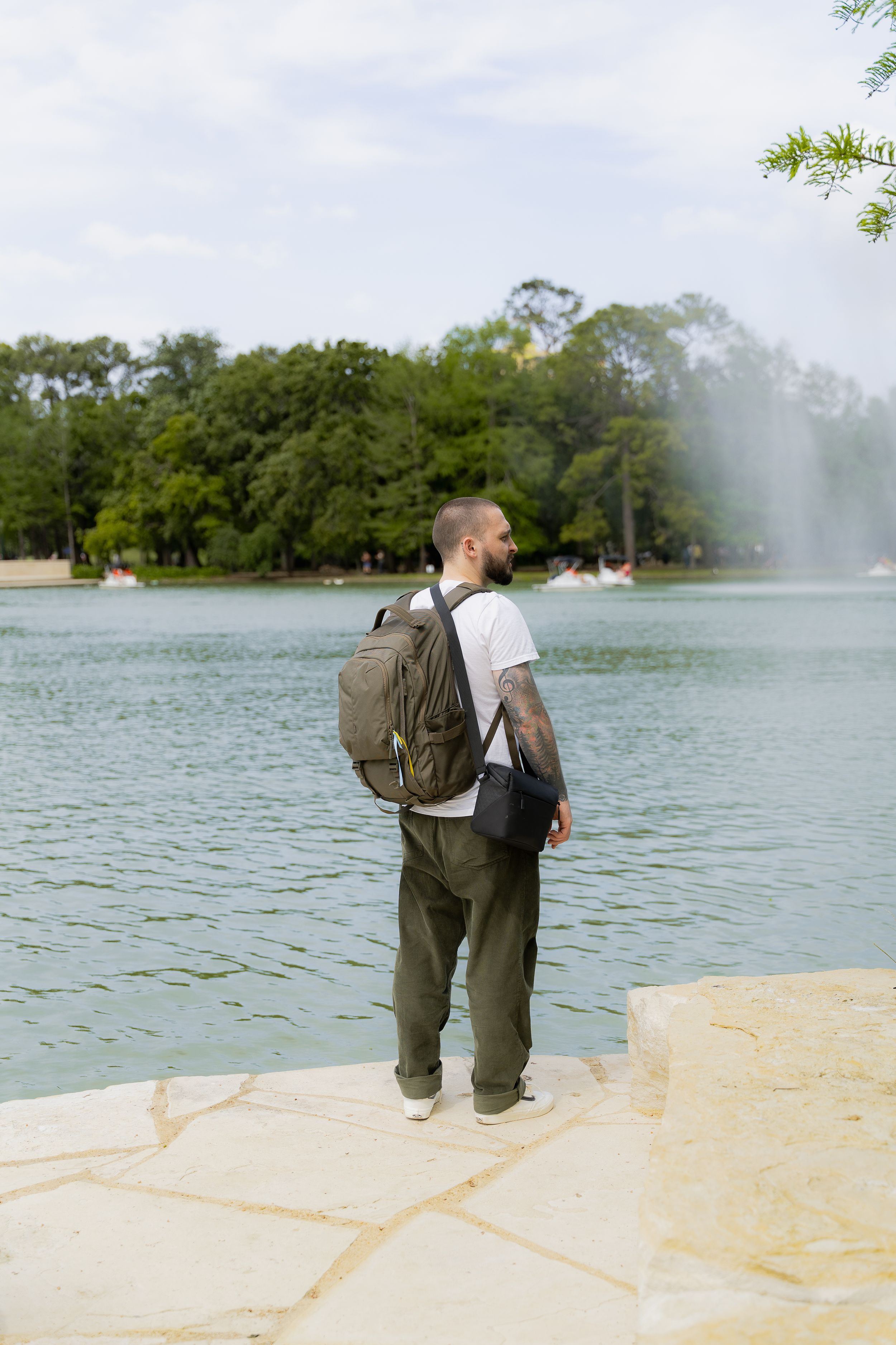 Person in casual attire with backpack stands at edge of lake with fountain and trees in background.
