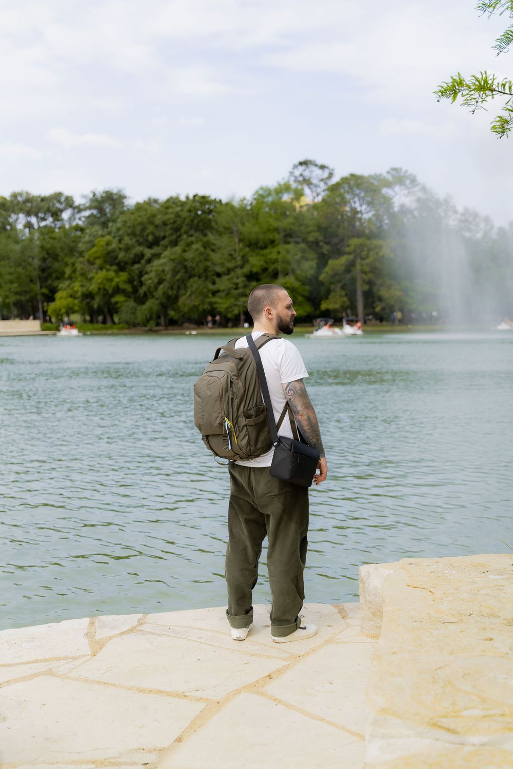 Person in casual attire with backpack stands at edge of lake with fountain and trees in background.